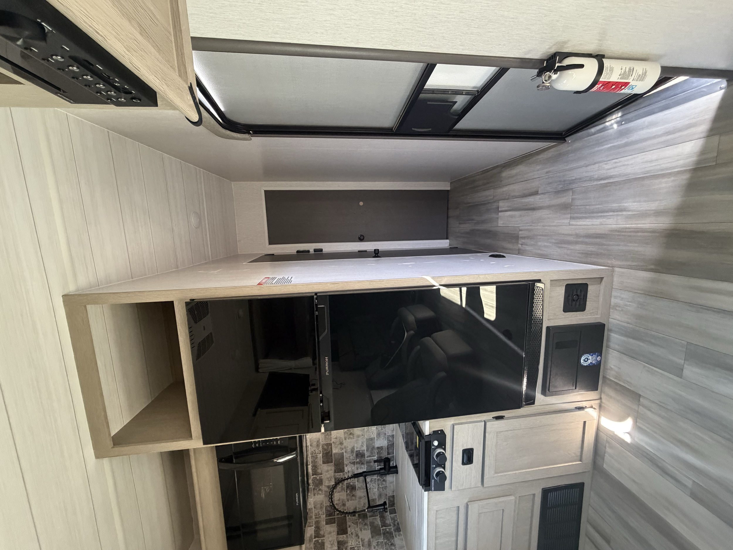 Interior of a small RV or camper, showing a kitchenette area with a microwave, stove, and refrigerator. There are wood cabinets and a fire extinguisher mounted on the ceiling. The floor is a light wood finish, and the walls are decorated in neutral t