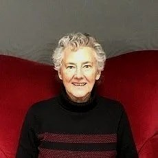Older woman with short gray hair smiling, wearing a black sweater, sitting on a red piece of furniture against a gray background.