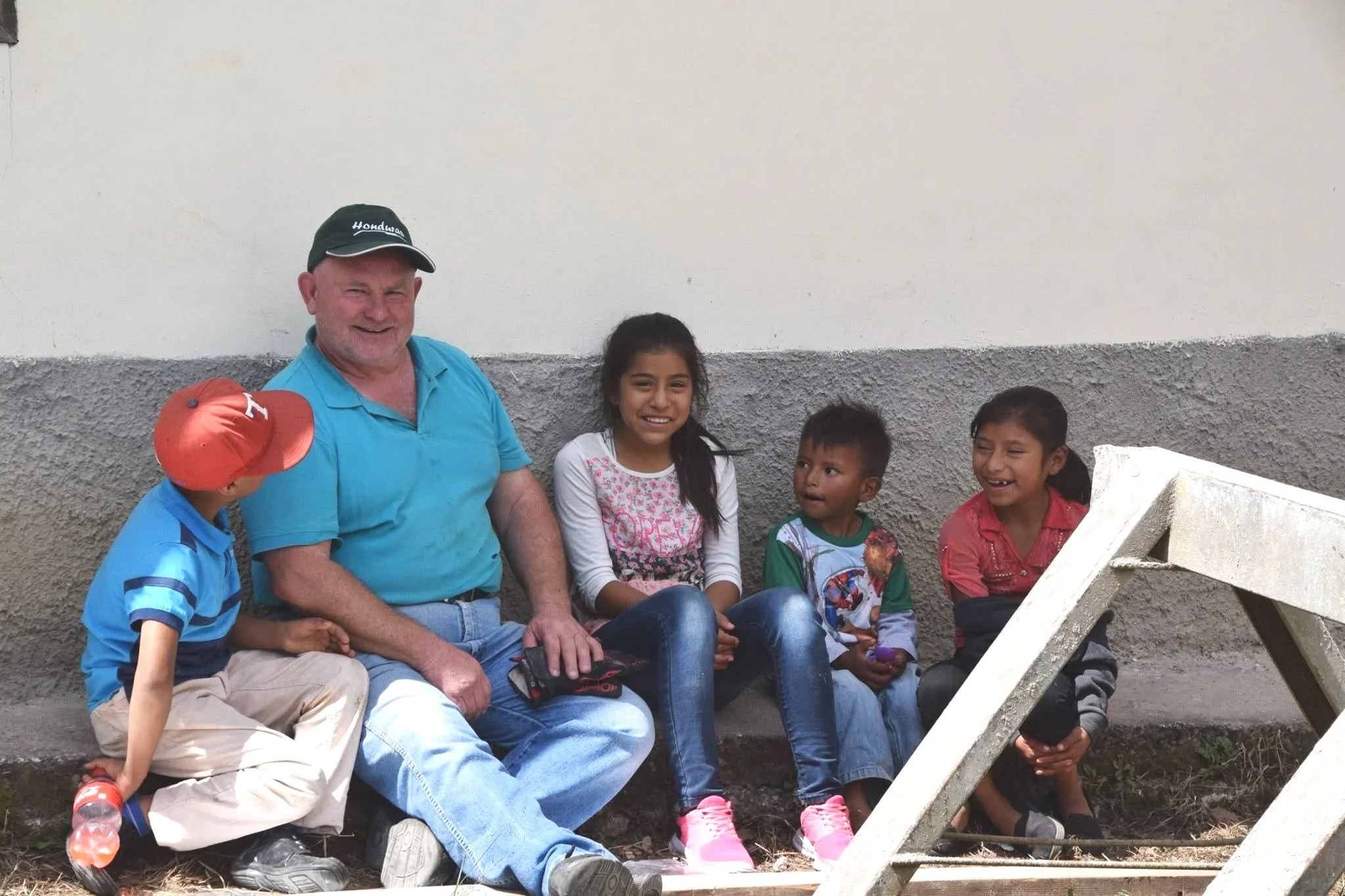 A group of five children and one adult man sitting on a bench against a two-tone concrete wall, smiling and enjoying each other's company. The man wears a black cap and a blue shirt. The children are dressed casually, with one wearing a hat and others in colorful clothing. One child is looking at a phone, while others look at the camera or each other.