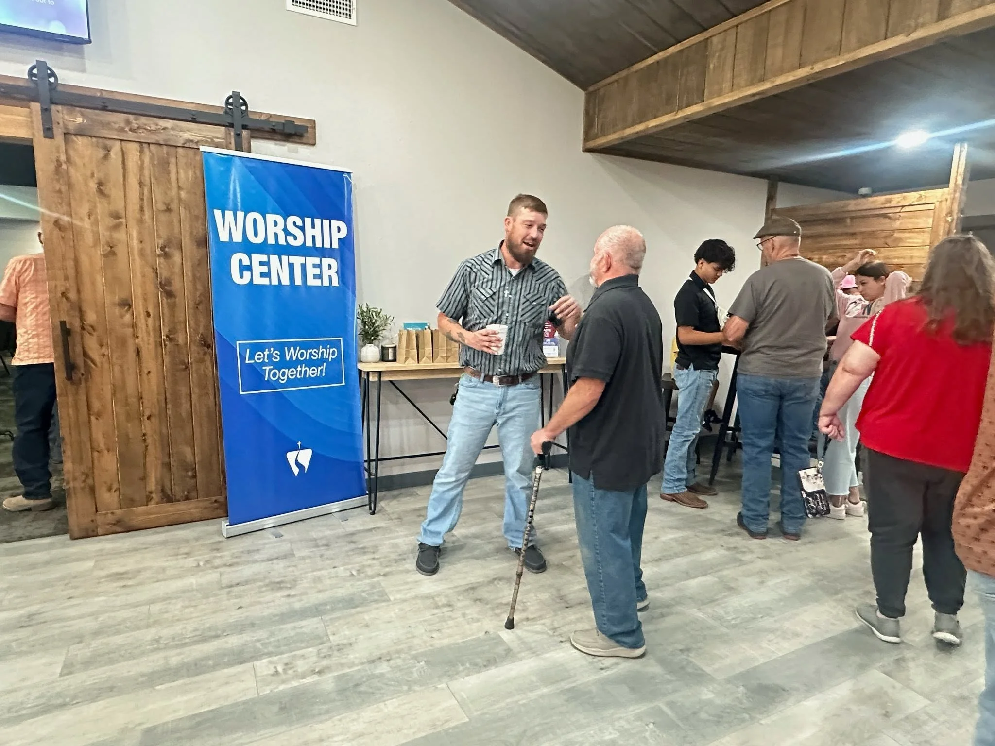 People gathered at a worship center, engaging in conversation and prayer, with a blue sign that reads 'WORSHIP CENTER' and a slogan 'Let's Worship Together.'
