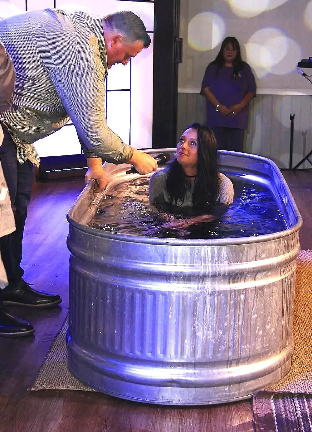Woman being baptized in a metal tub filled with water, with a man holding a microphone or speaker and speaking to her, and a woman standing in the background watching.