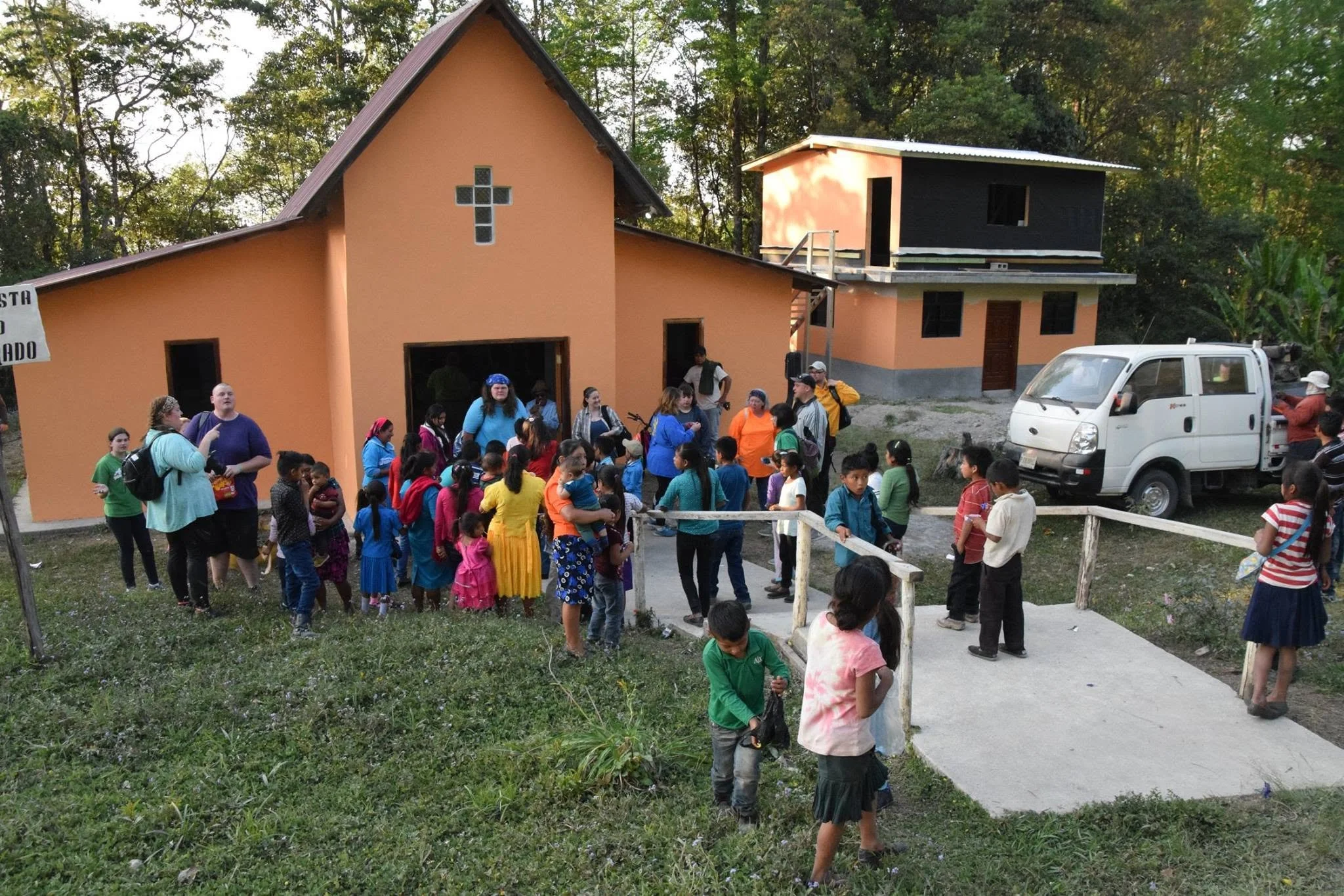 A group of children and adults gather outside a church with a peach-colored exterior and a small cross on the front wall, surrounded by trees. Some children are wearing colorful clothing, and some adults are talking or supervising. A small white van is parked nearby.