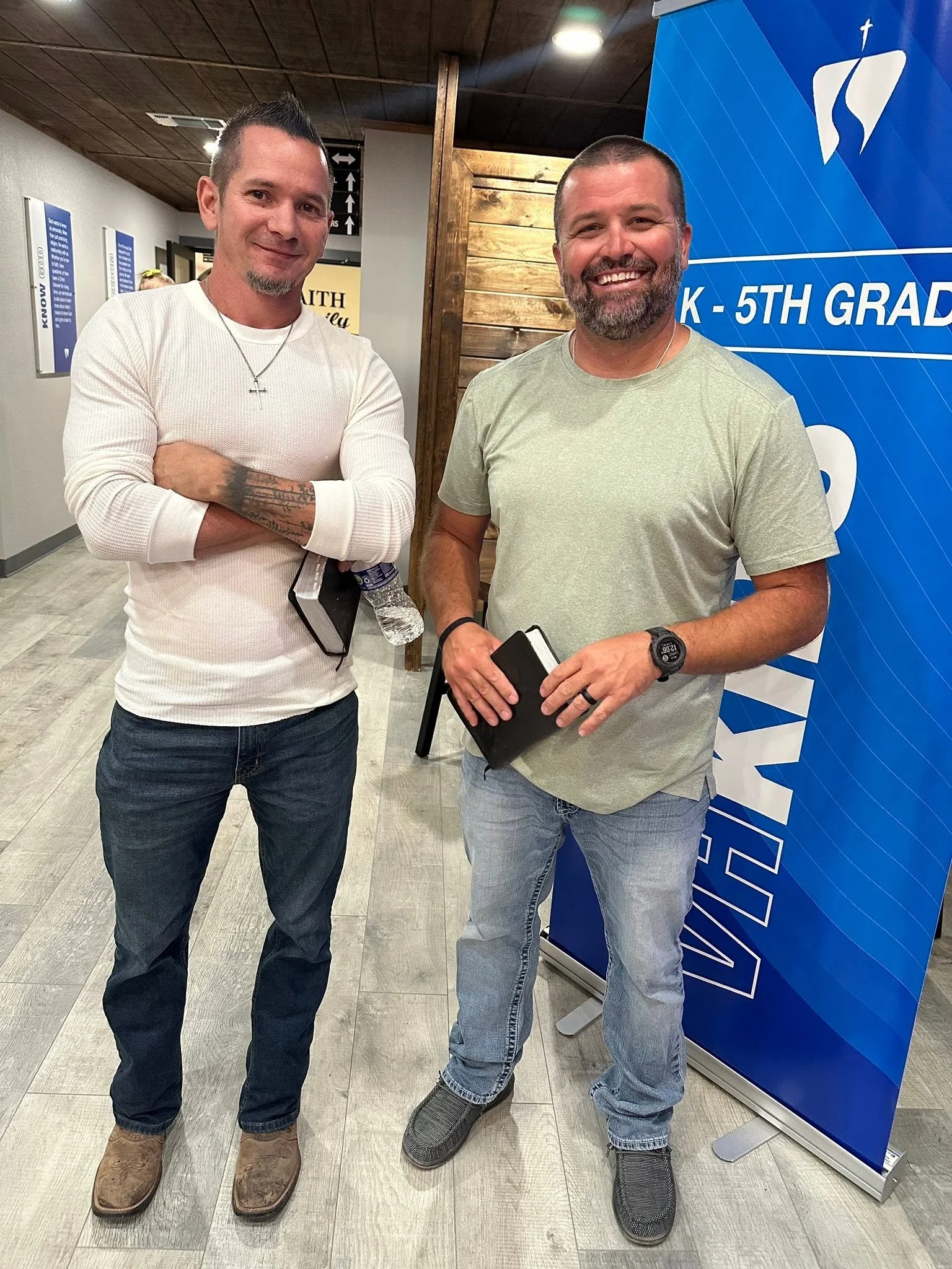 Two men standing indoors, smiling at the camera. One has short dark hair, a goatee, a cross necklace, and is wearing a white long-sleeve shirt, dark jeans, and brown boots. The other has short dark hair, a beard, and is wearing a light gray t-shirt, gray jeans, and slip-on shoes. They are standing in front of a blue banner with white text and graphics, and the background includes a wooden wall and some posters.