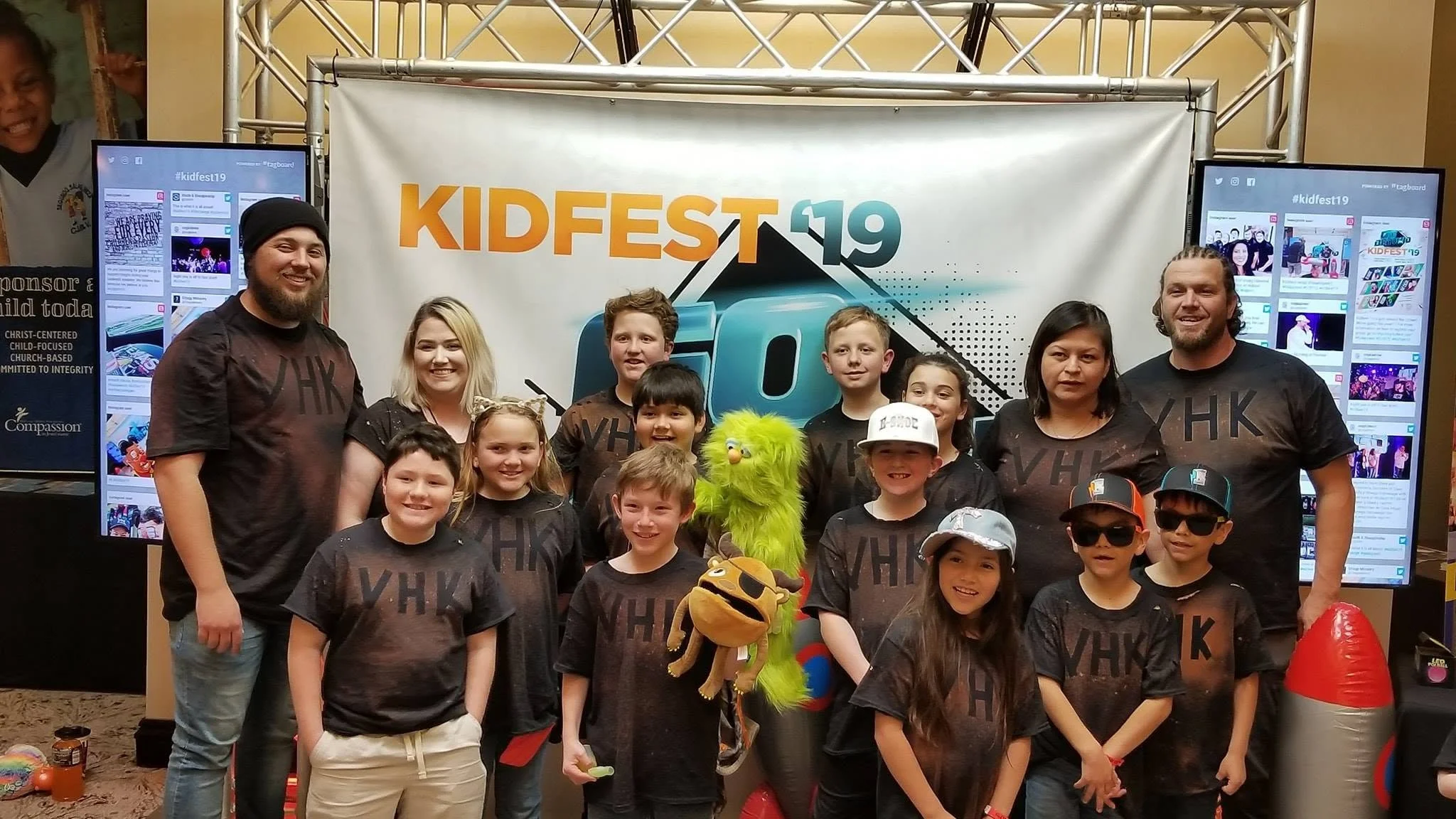 Group photo of children and adults at a Kidsfest event, standing in front of a banner with the event's name. Kids are smiling, some wearing sunglasses, holding toys and balloons, with a mascot in the center.