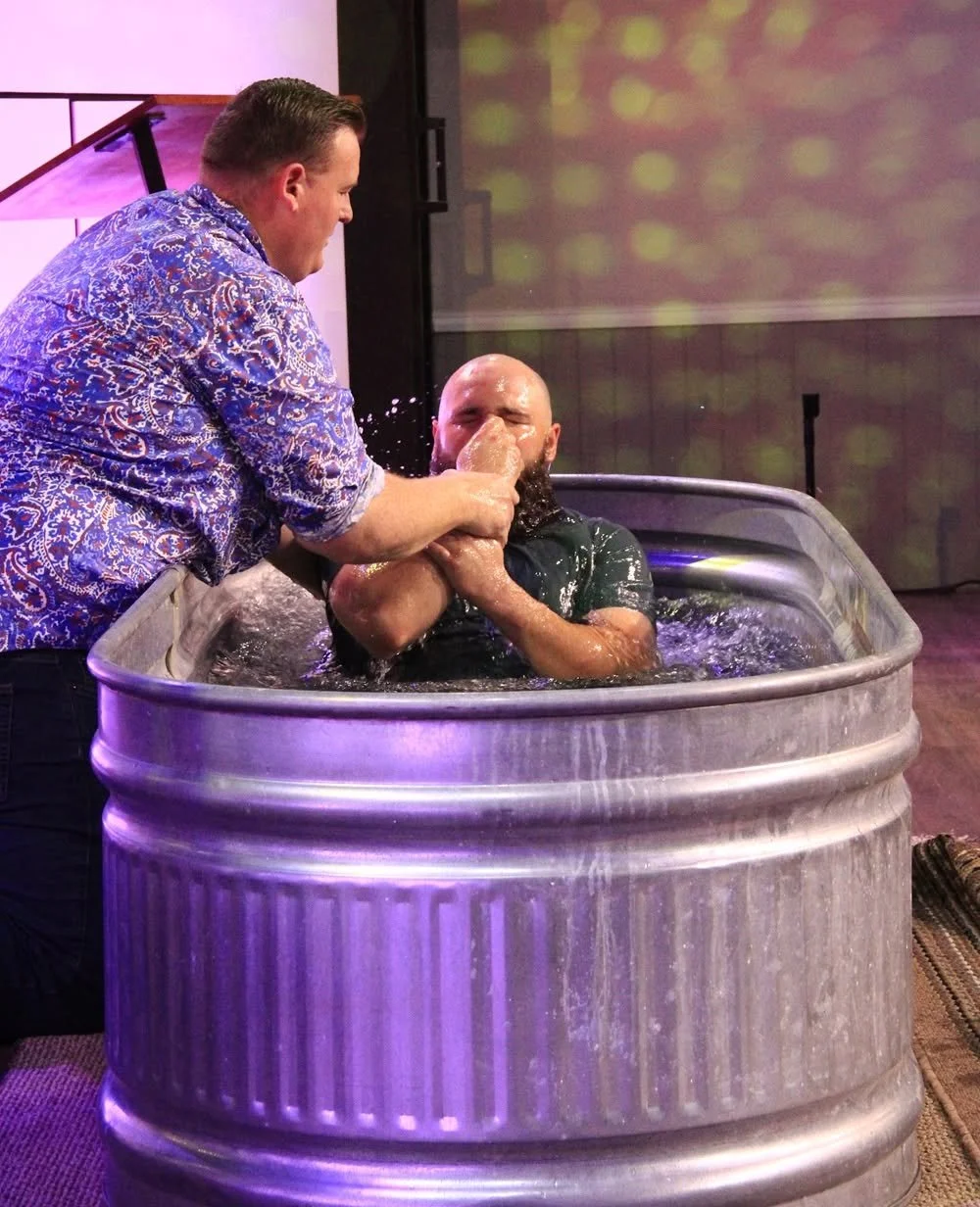 Two men engaged in a baptism ceremony, with one man submerged in a tub filled with water and the other man holding his nose, preparing for immersion. The setting appears to be indoors with purple and yellow lighting.