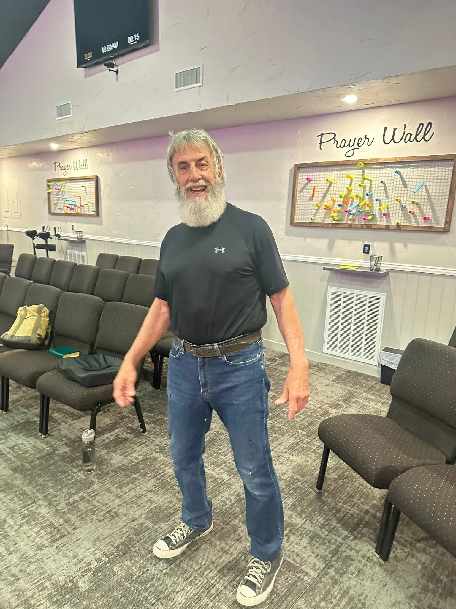 An older man with a gray beard and gray hair smiling, wearing a black Under Armour t-shirt, blue jeans, and black and white Converse sneakers, standing in a church or community room with rows of black chairs, a water bottle, and a backpack nearby.