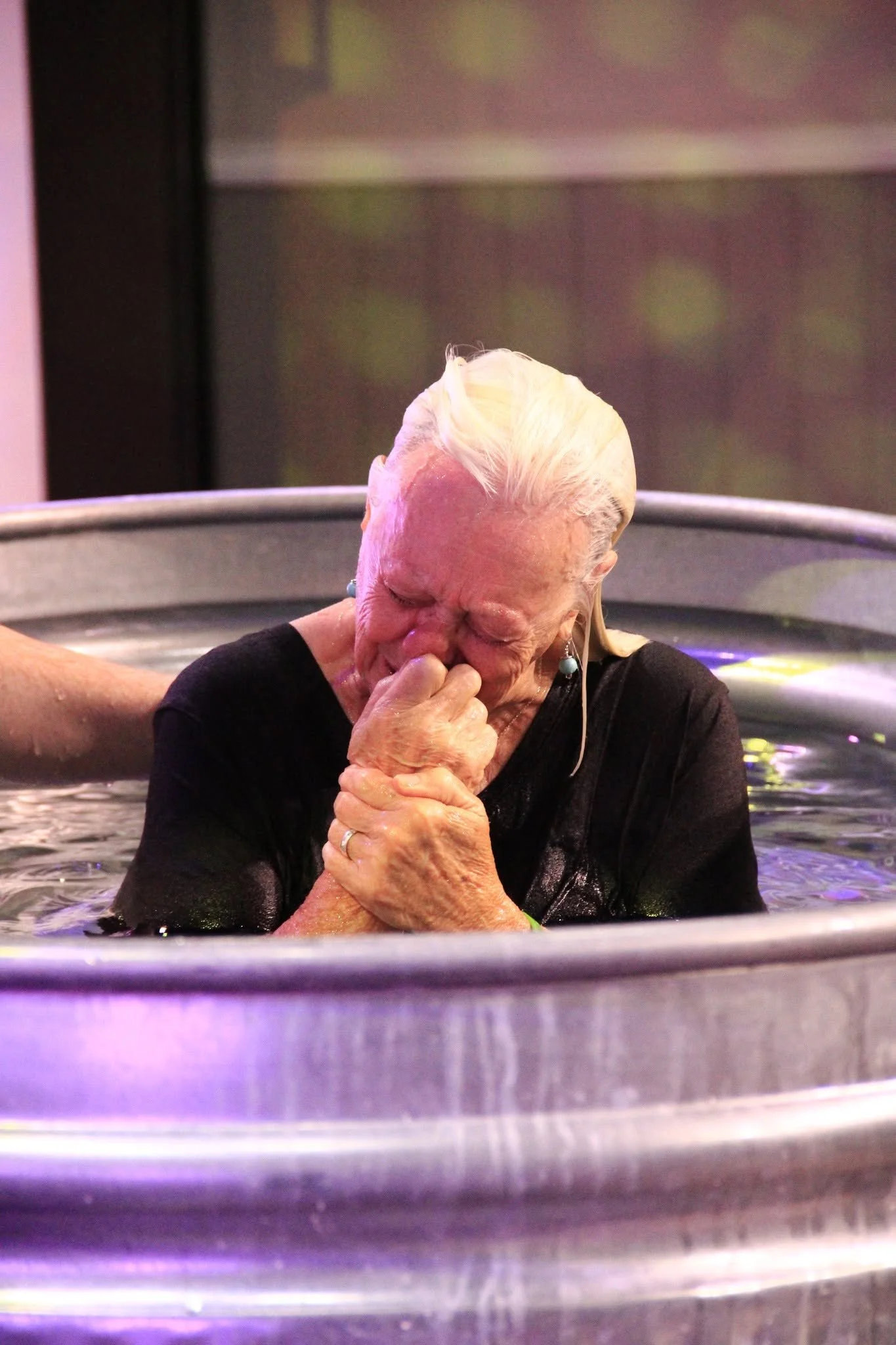 An elderly woman with white hair, wearing a black top, is baptized in a baptismal pool, with her eyes closed and hands clasped together, appearing emotional.
