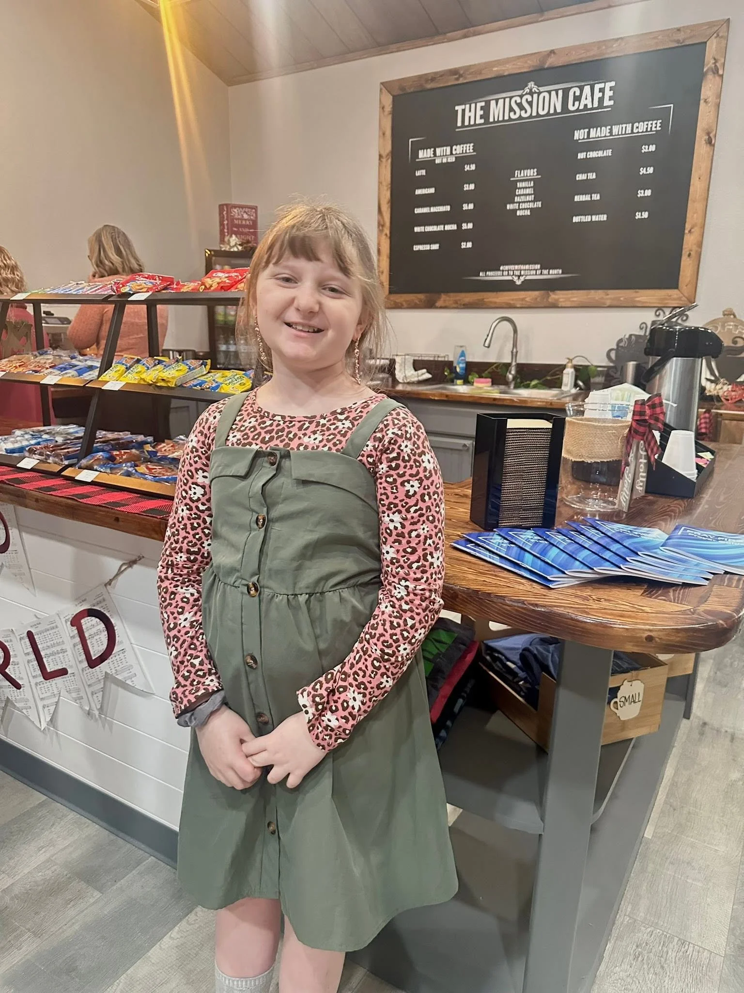A young girl with light brown hair smiling at the camera, standing in a cafe with a blackboard menu in the background. She is wearing a pink leopard-print long-sleeve shirt and a green dress with buttons. Behind her, there is a display of packaged snacks and a counter with various items.