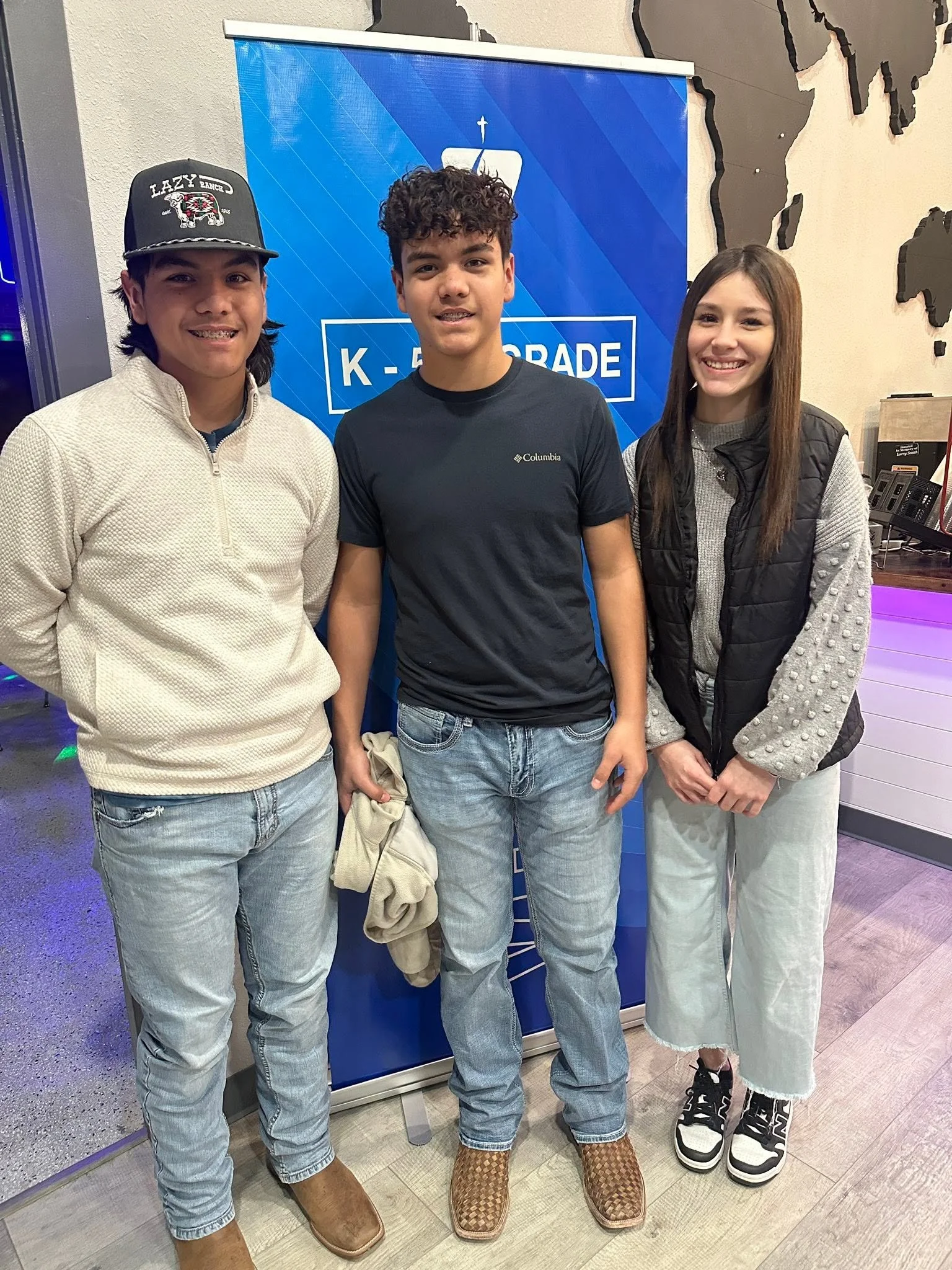 Three teenagers standing together indoors in front of a blue donation box. The boy on the left is wearing a black cap with a cow patch and the words 'Lazy Ranch', a beige zip-up sweater, and jeans. The boy in the middle has curly hair, is wearing a black Columbia t-shirt and jeans, holding a beige hoodie and wearing woven slippers. The girl on the right has straight, brown hair, is wearing a gray top, a black puffer vest, a beige skirt, and black-and-white sneakers. They are smiling at the camera.