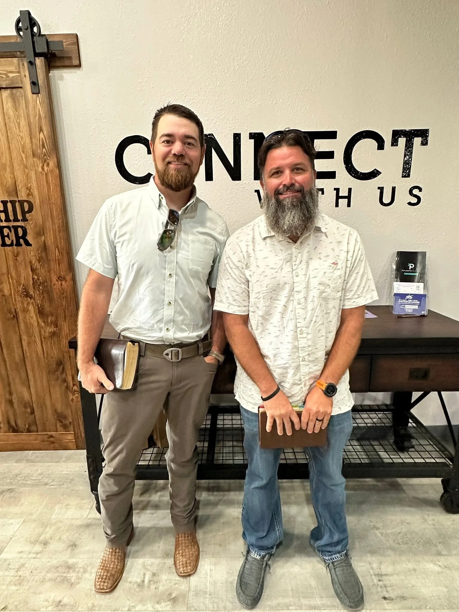 Two men standing indoors in front of a wall with the words 'CONNECT with us'. The man on the left wears a white shirt, khaki pants, and patterned shoes, holding a book. The man on the right has a beard, wears a light-colored patterned shirt, jeans, and gray shoes, holding a book. There are pamphlets on a table behind them.