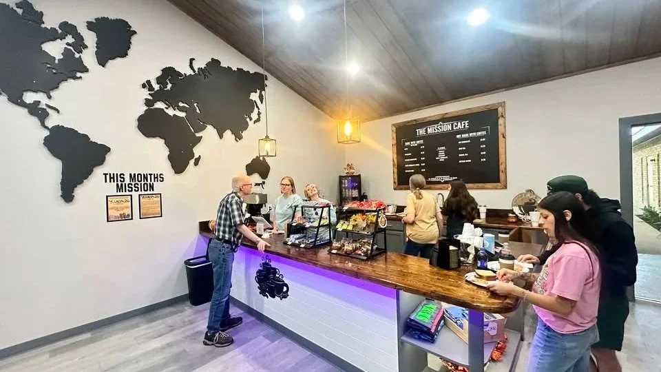 Inside a cafe with a large world map on the wall and a blackboard menu titled 'The Mission Cafe.' Customers are ordering at the counter, and staff are serving behind it. There are purple neon lights under the counter and framed mission-related posters on the wall.