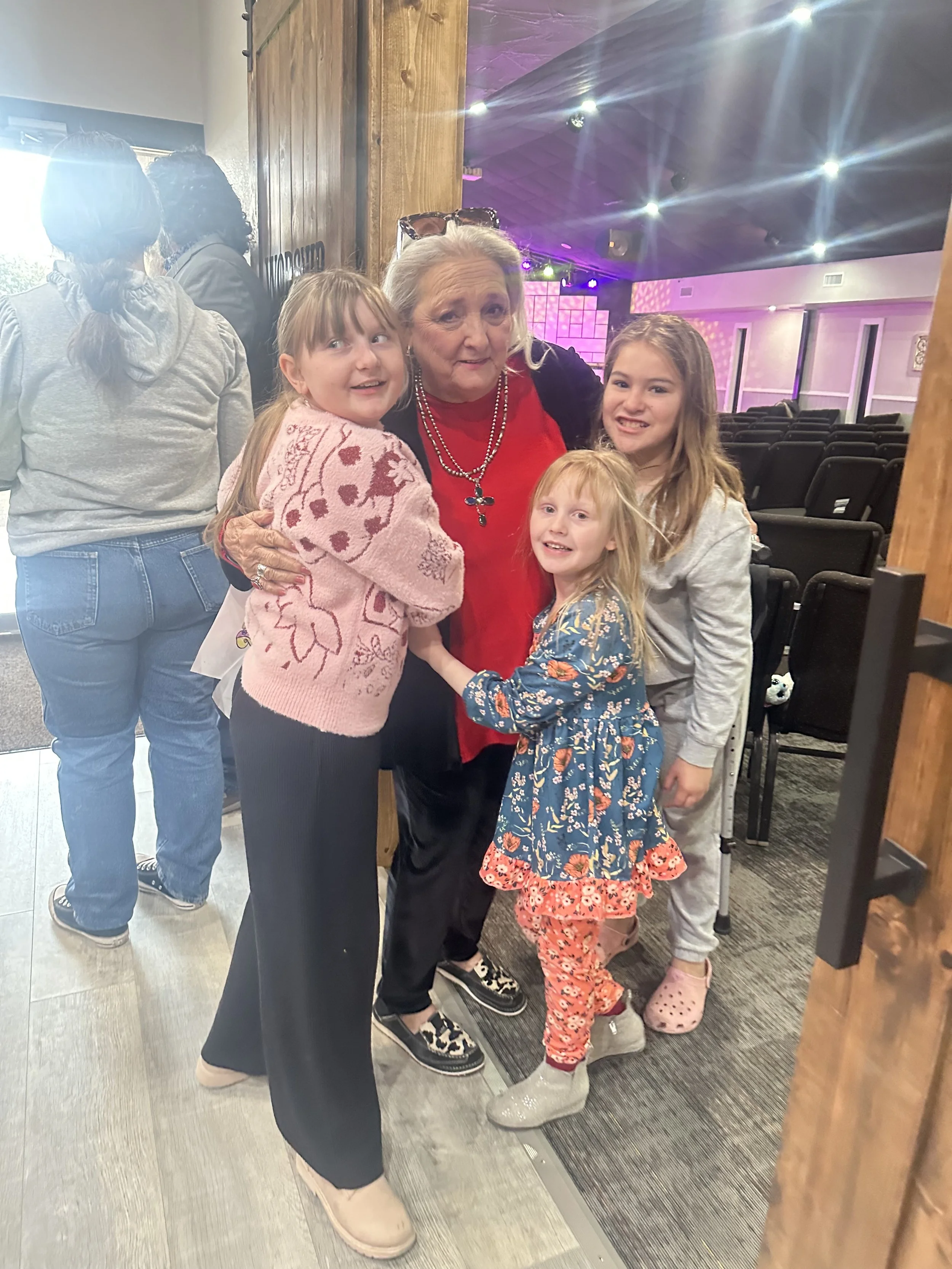 A group of children and an elderly woman are smiling and posing together in a room with theater-style seating and colorful lighting in the background. The children are hugging the elderly woman and looking happy.