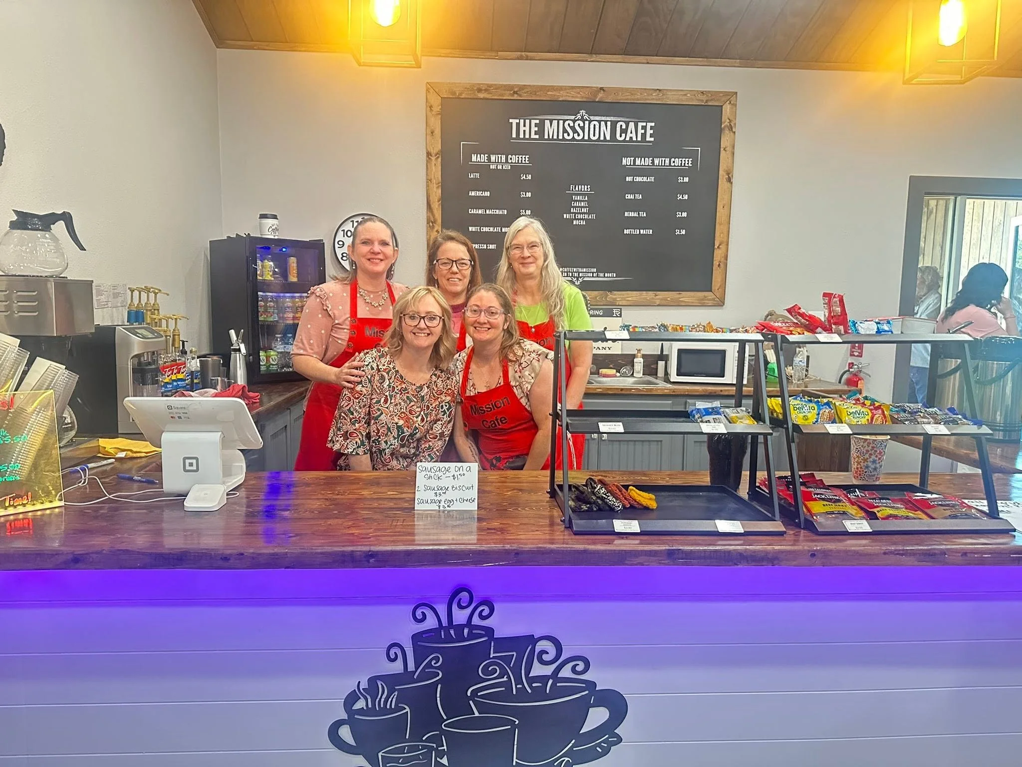 Four women behind the counter at The Mission Cafe, smiling and wearing red aprons. The cafe menu is on the wall behind them, with coffee and non coffee drink options. The counter displays various snacks, a card payment device, and a handwritten sign listing sausage options and prices.