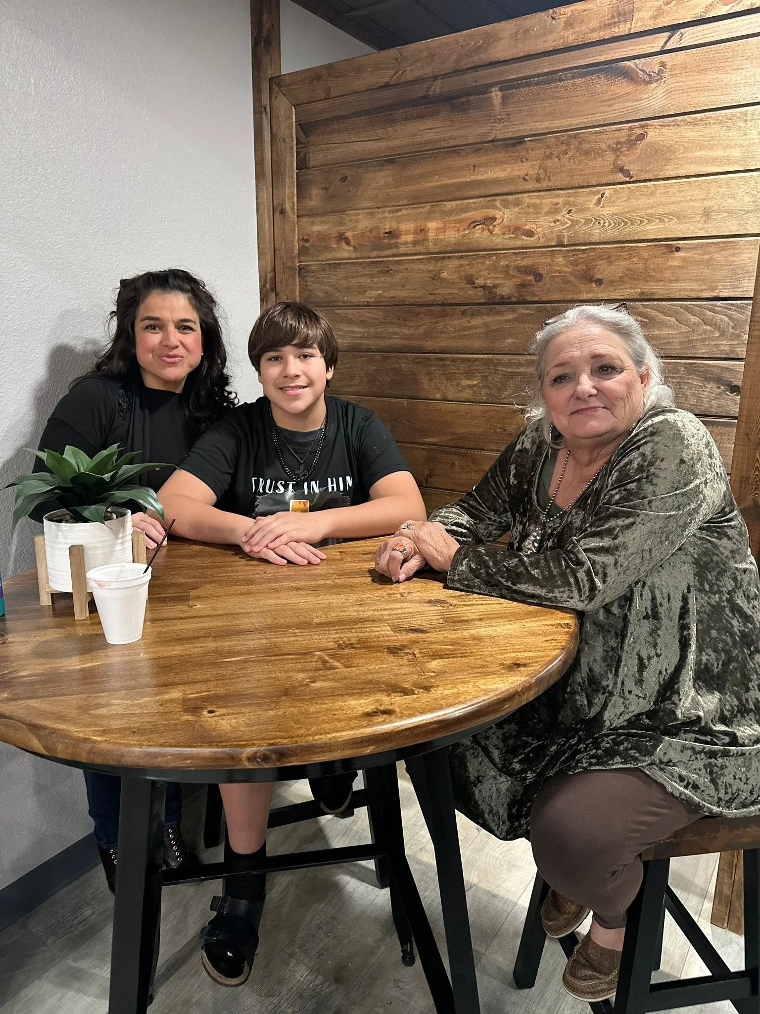 Three people, two women and one young man, sitting at a wooden table in a cafe. One woman has long dark hair, the young man has short brown hair, and the older woman has gray hair. There is a green potted plant and a drink on the table.