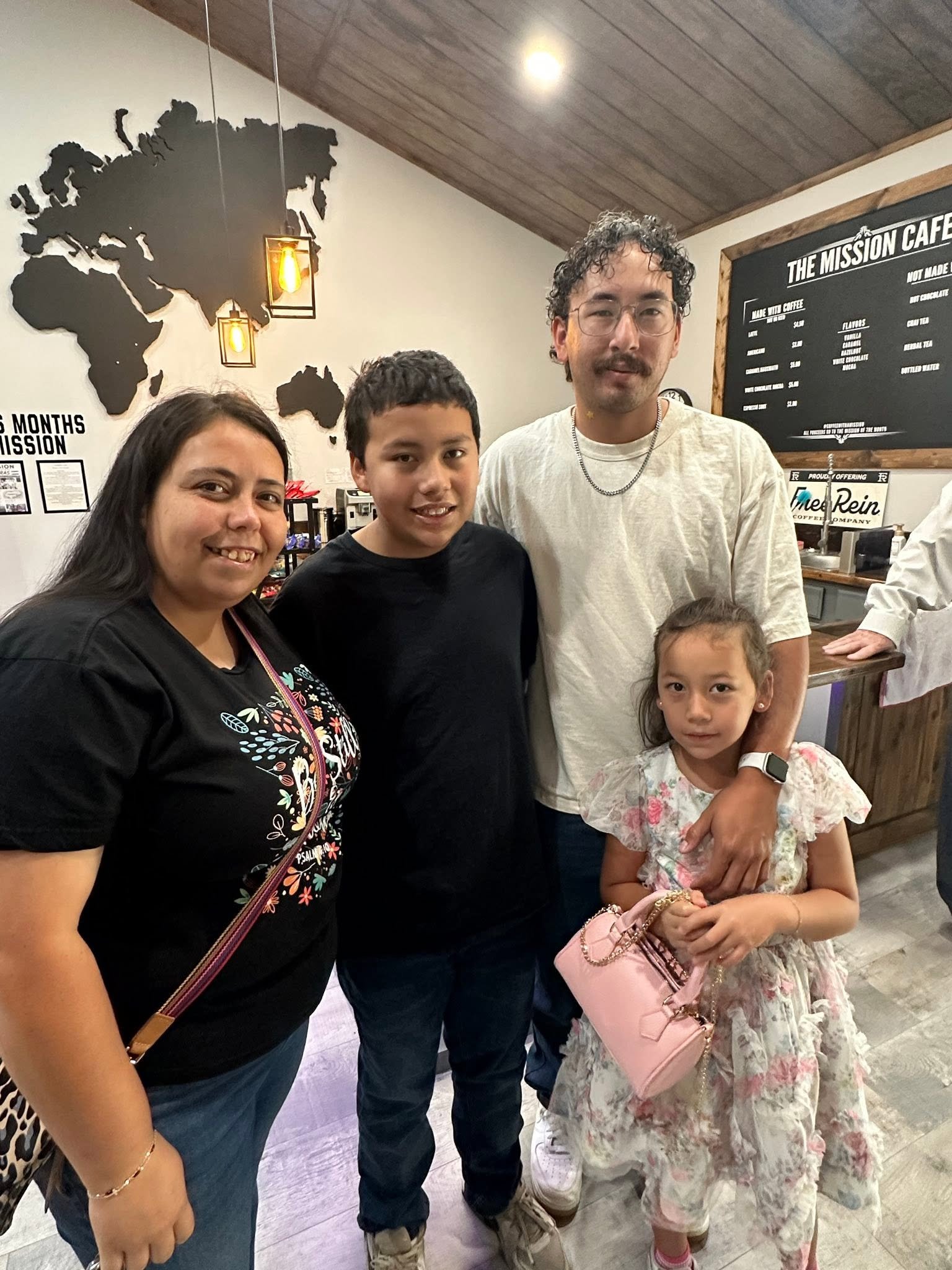 Group of four people, three adults and one child, indoors at a cafe or restaurant. They are smiling and standing close together. The woman on the left has dark hair, the young boy has short dark hair, the man on the right has curly hair and glasses, and the young girl has long hair and is holding a pink bag.
