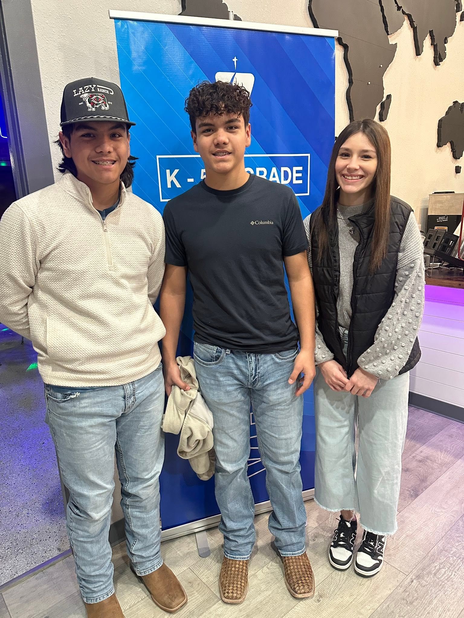 Three teenagers are standing indoors in front of a blue sign that says 'K-12 GRADE'. The boy on the left is wearing a baseball cap, a cream sweater, and jeans. The boy in the middle is wearing a black Columbia t-shirt, jeans, and patterned slippers. The girl on the right has long brown hair, a gray sweater under a black vest, and light-colored pants with black and white sneakers.