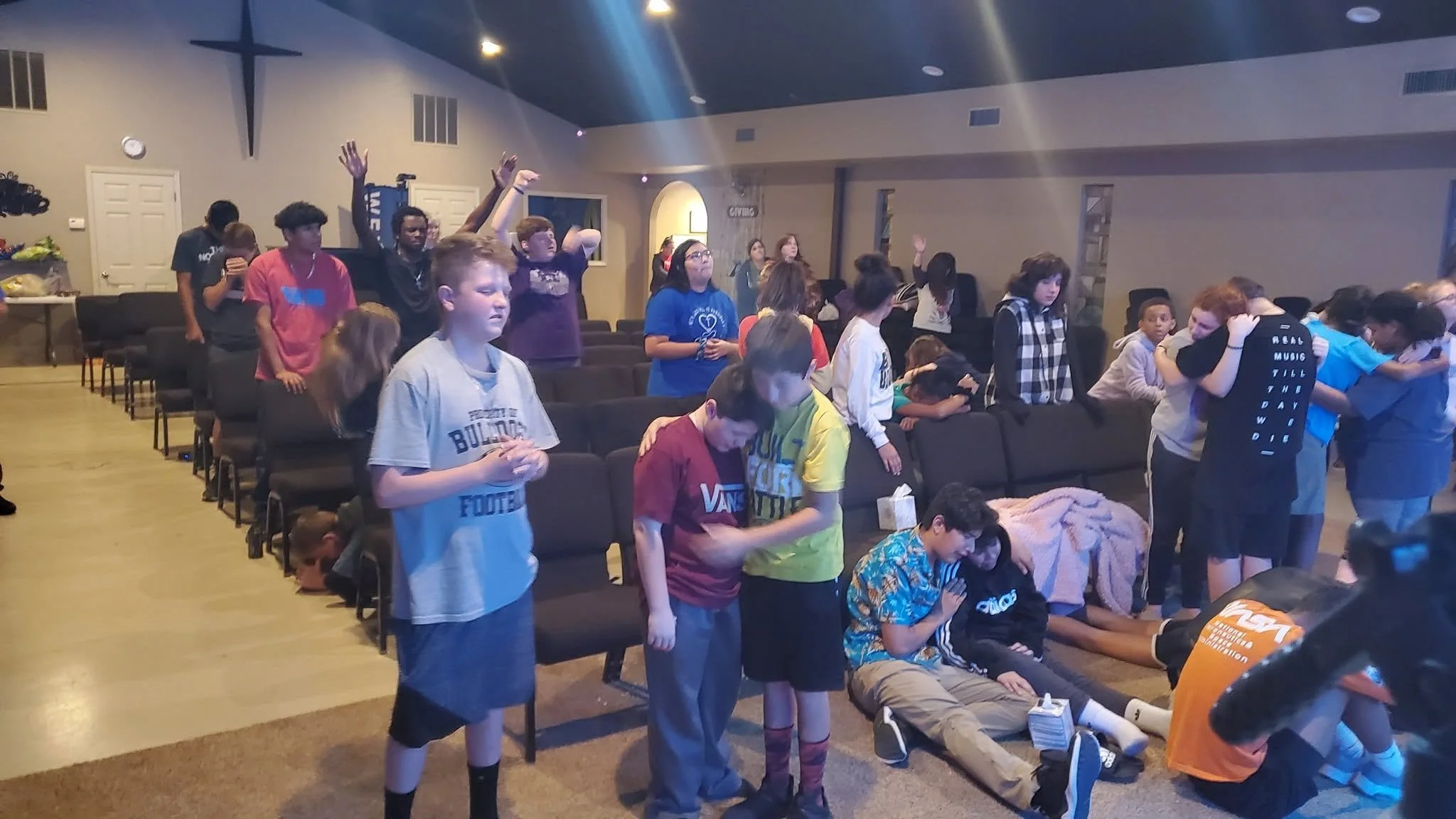 Children and teenagers gather in a church or community room, some prayerfully with heads bowed, others standing or sitting, engaged in a group activity or prayer.