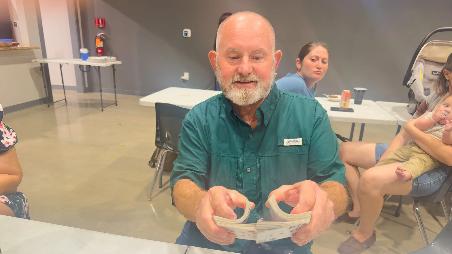 A man with a white beard, wearing a green shirt with a name tag that reads 'Magellan', sitting at a table and holding playing cards, with people and a high chair in the background.
