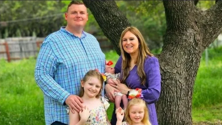 Family of five outdoors under a large tree, with a man, woman, two young girls, and a baby in the woman's arms.