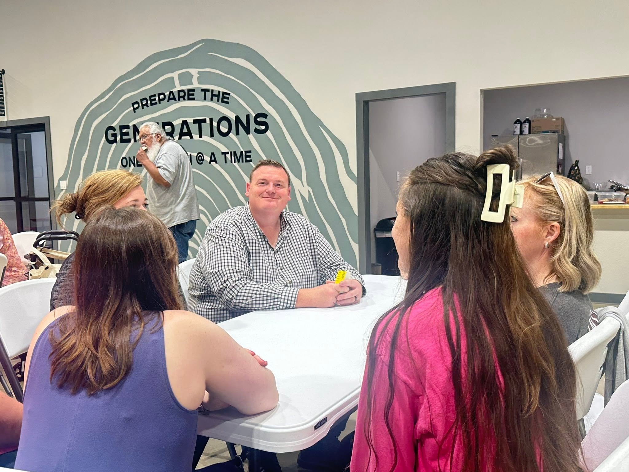 A group of people sitting around a table during a gathering or meeting, with a mural on the wall behind them that says 'Prepare the Generations One at a Time'.