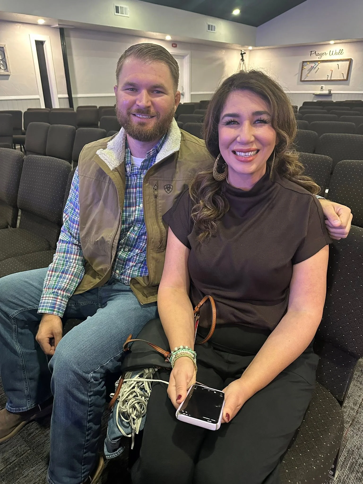 A man and a woman sitting close together in a church or auditorium, smiling at the camera. The man has short hair, a beard, and is wearing a plaid shirt with a tan vest. The woman has long, wavy hair, and is wearing a dark blouse with large earrings. She holds a smartphone in her lap, and she is wearing several colorful bracelets on her left wrist.