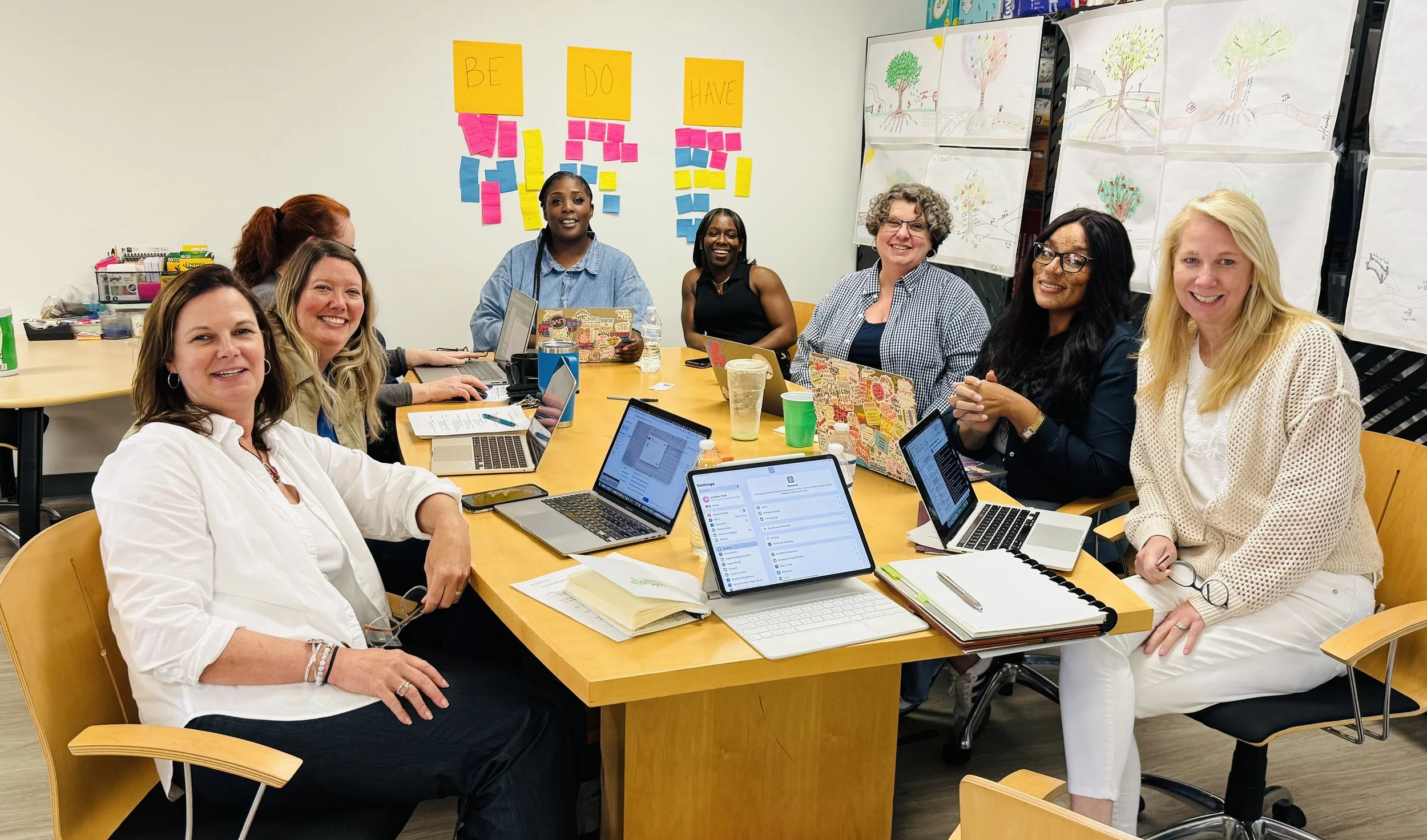 A diverse group of nine women gathered around a large conference table in a meeting room. They are smiling and working on laptops, with colorful sticky notes and drawings of trees on the walls behind them.
