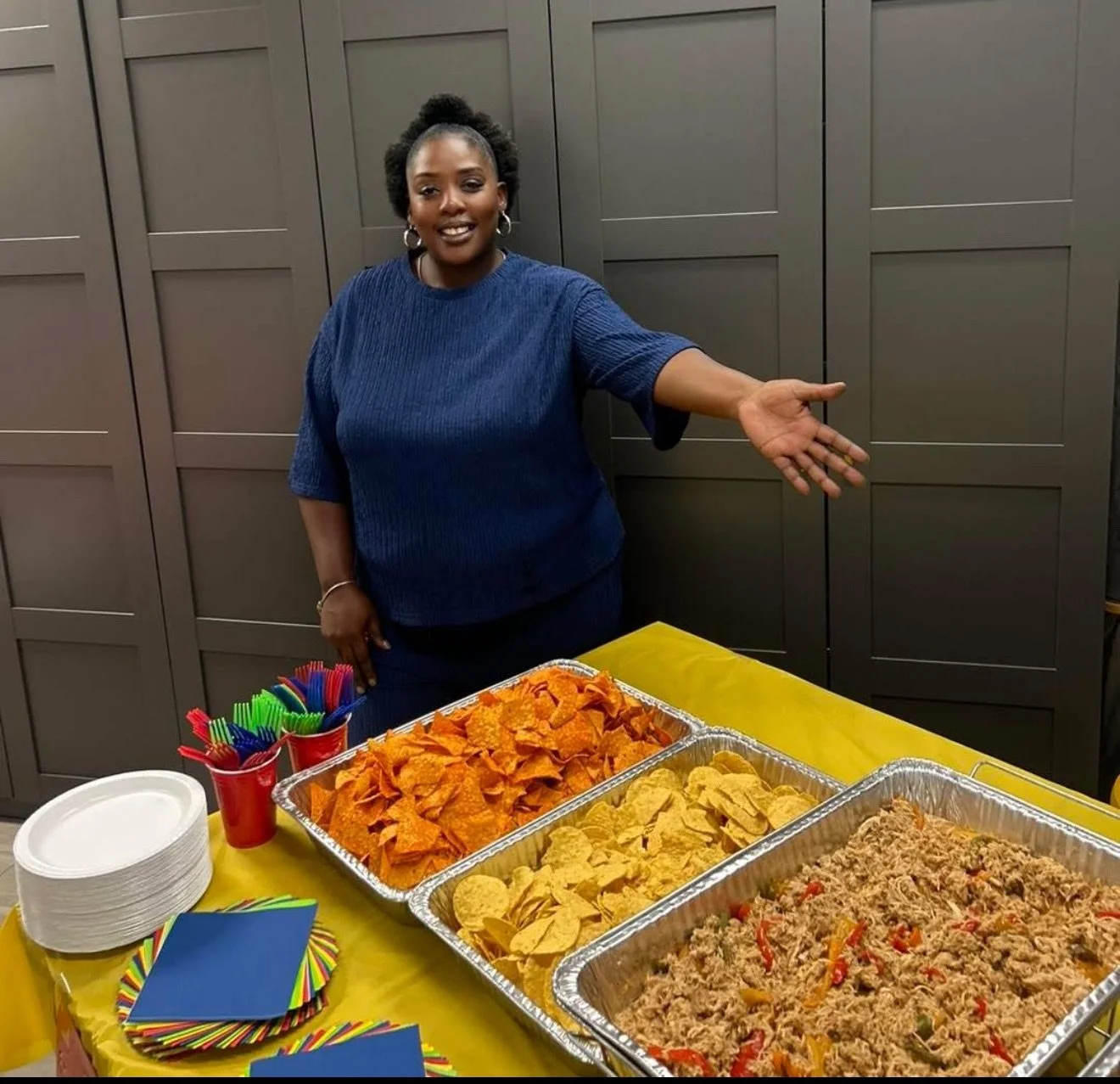 Woman standing behind a table with trays of chips, dip, and taco meat, with colorful straws, plates, and napkins on the table.