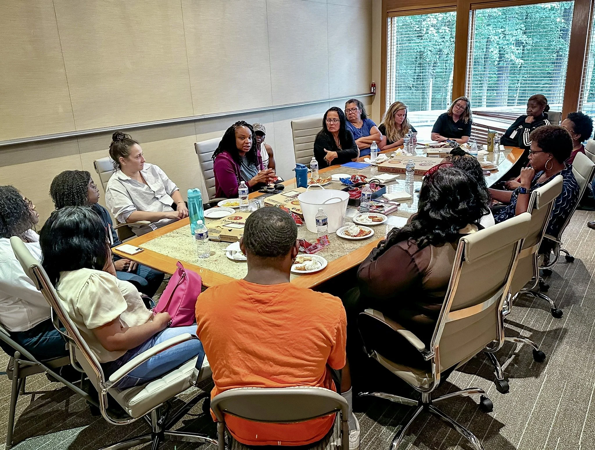 A diverse group of people sitting around a large conference table during a meeting, with pizza boxes, plates, water bottles, and snacks on the table, in a room with large windows and natural light.