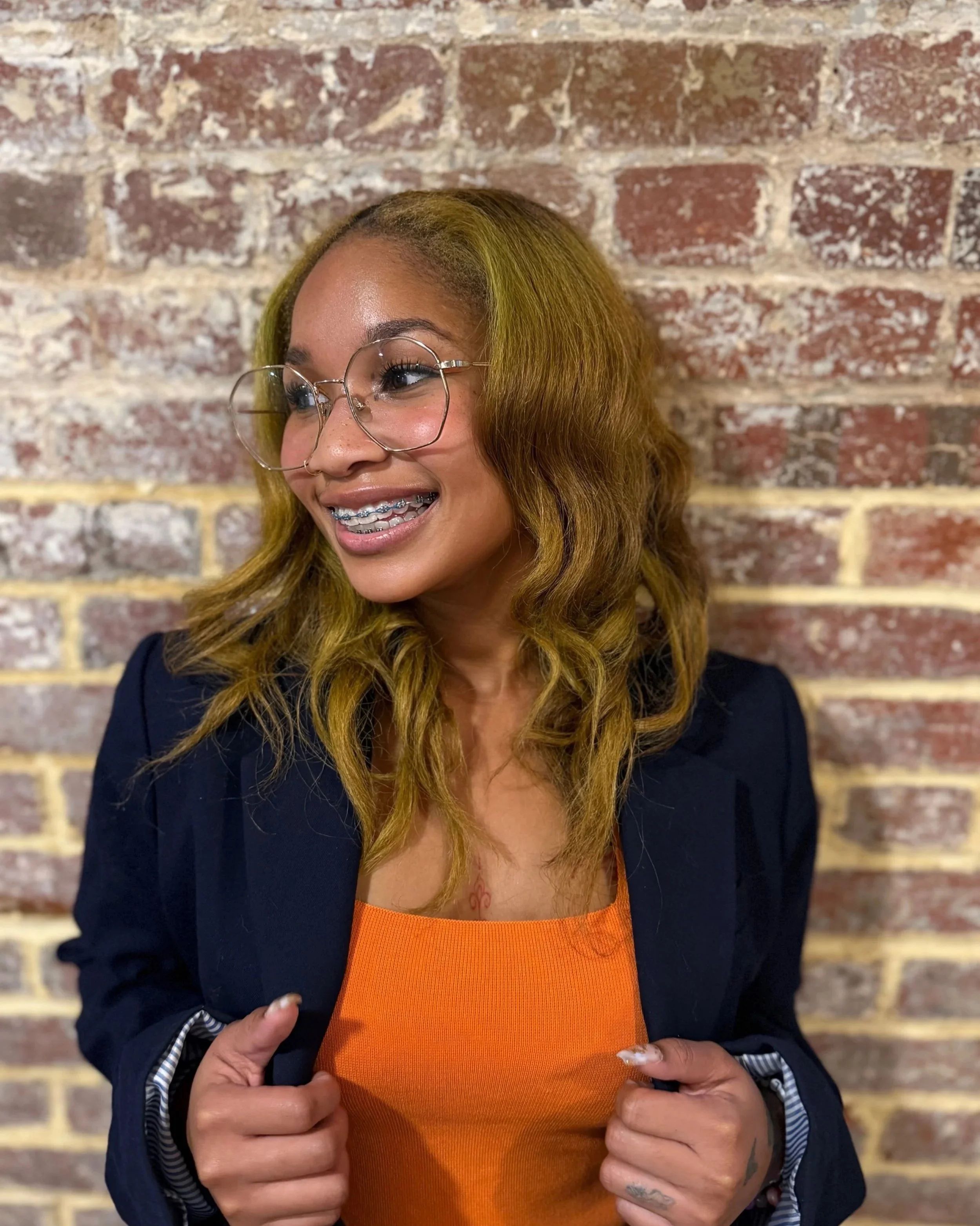 A smiling woman with long, wavy, light brown hair wearing glasses, a black blazer, and an orange top, standing against a brick wall.