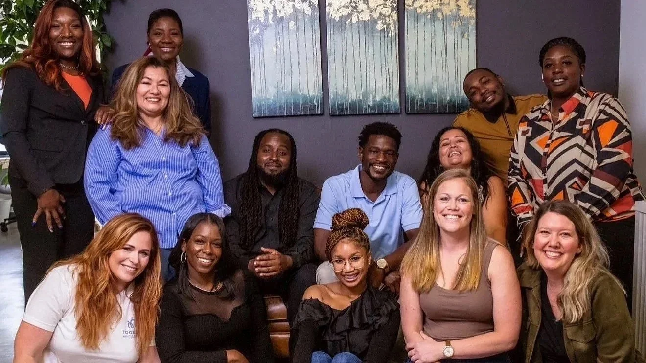 Group of eleven diverse women and men smiling and posing indoors, some standing and some sitting, against a dark wall with abstract art and a plant in the background.