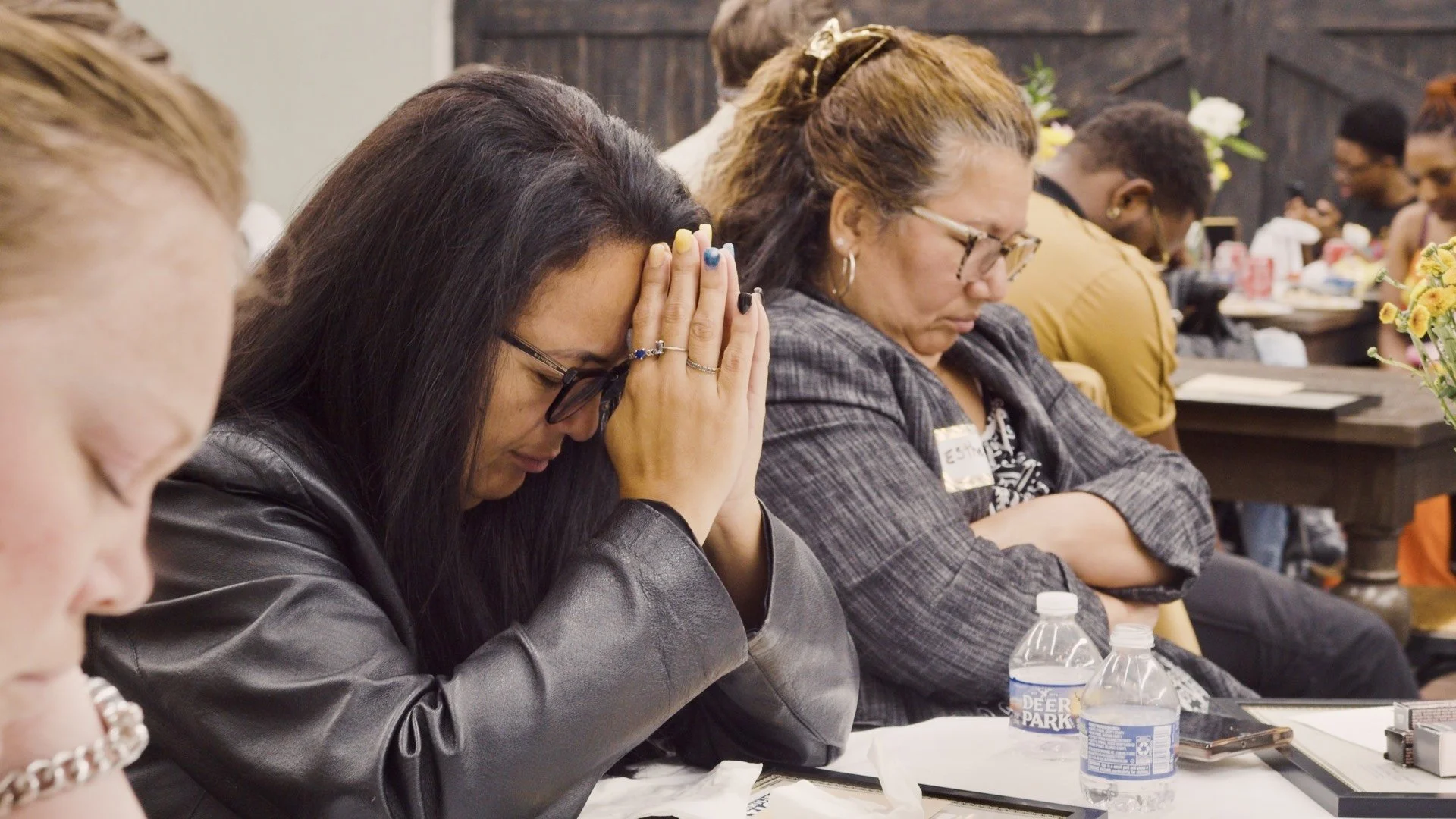 Group of people praying or meditating with heads bowed and hands clasped in a conference or meeting room.