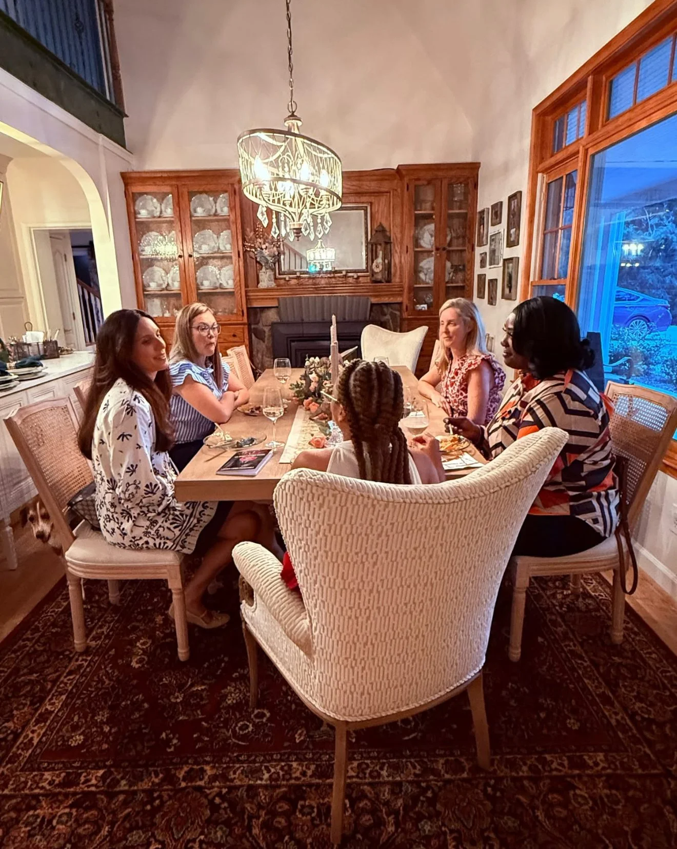 Six women gathered around a dining table in a cozy, well-lit dining room with wooden cabinets, a chandelier, and a large window showing the evening outside.