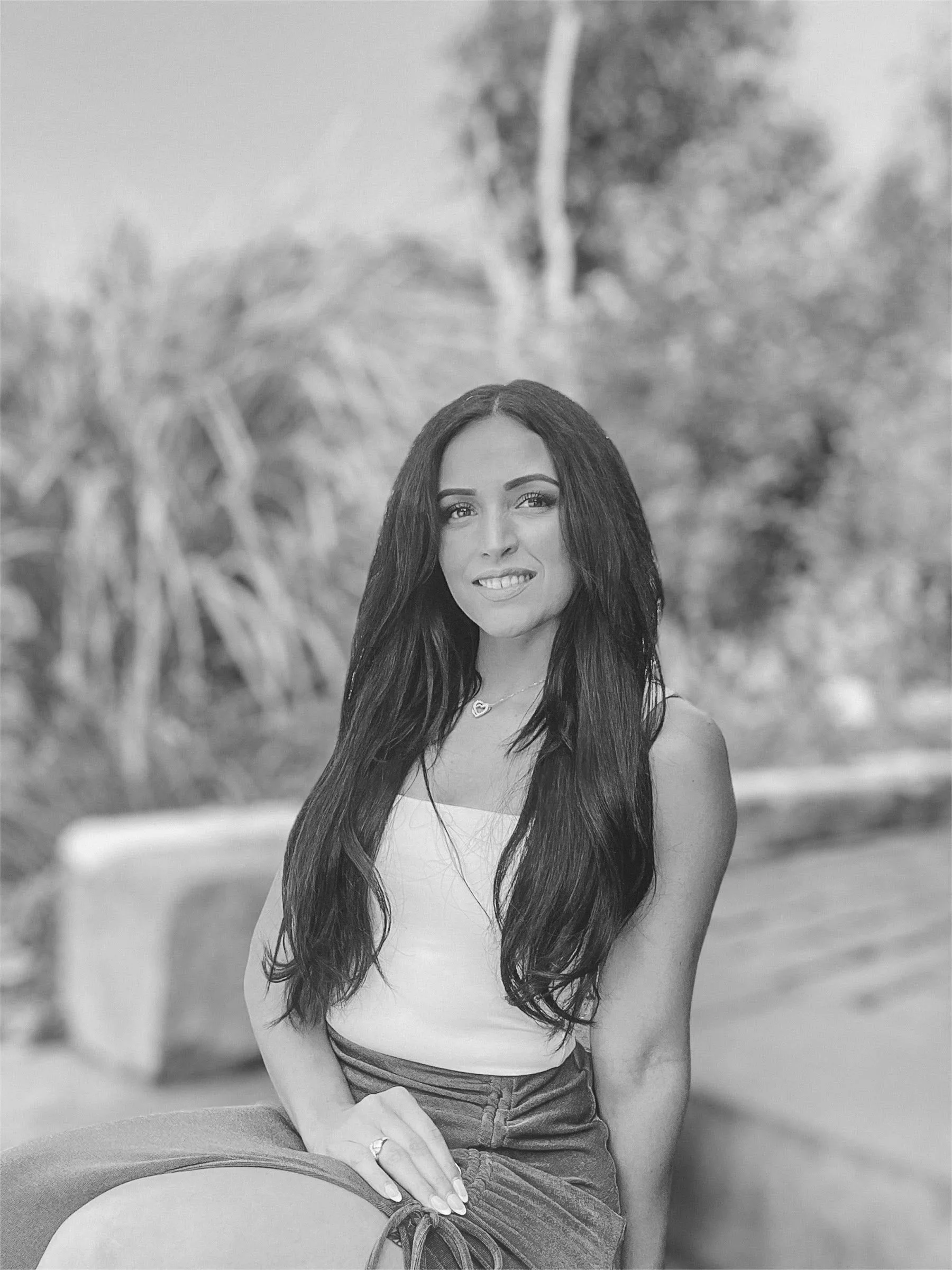 A black and white photo of a young woman with long dark hair, sitting outdoors on a stone bench, wearing a sleeveless top and a skirt, smiling at the camera.