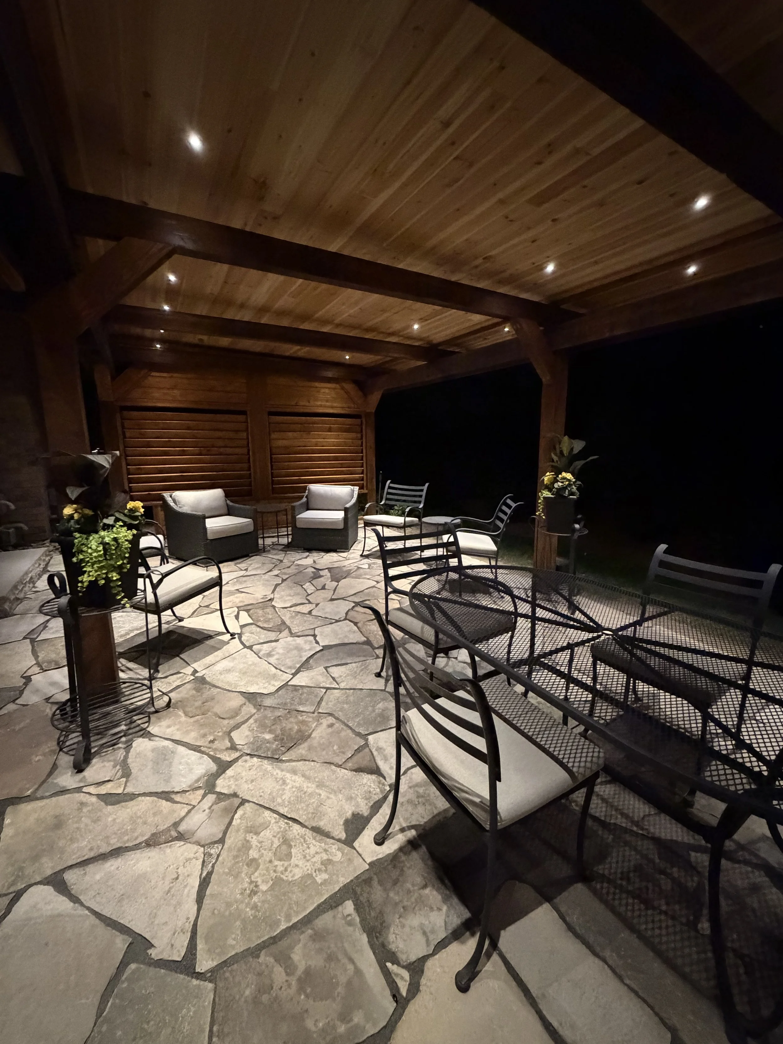 A backyard patio with a wooden pergola, outdoor furniture, potted plants, and a brick house with multiple rooflines and windows, surrounded by lush greenery and colorful bushes.