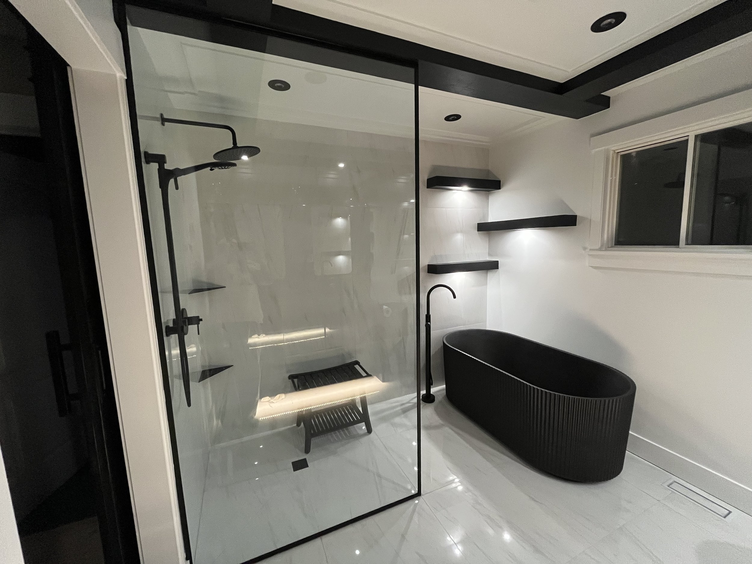 Modern bathroom featuring a white vanity with a marble countertop, black faucet, and black accessories, a round wall mirror, wall-mounted light fixture, a small window, framed artwork, a potted plant, and a toilet.