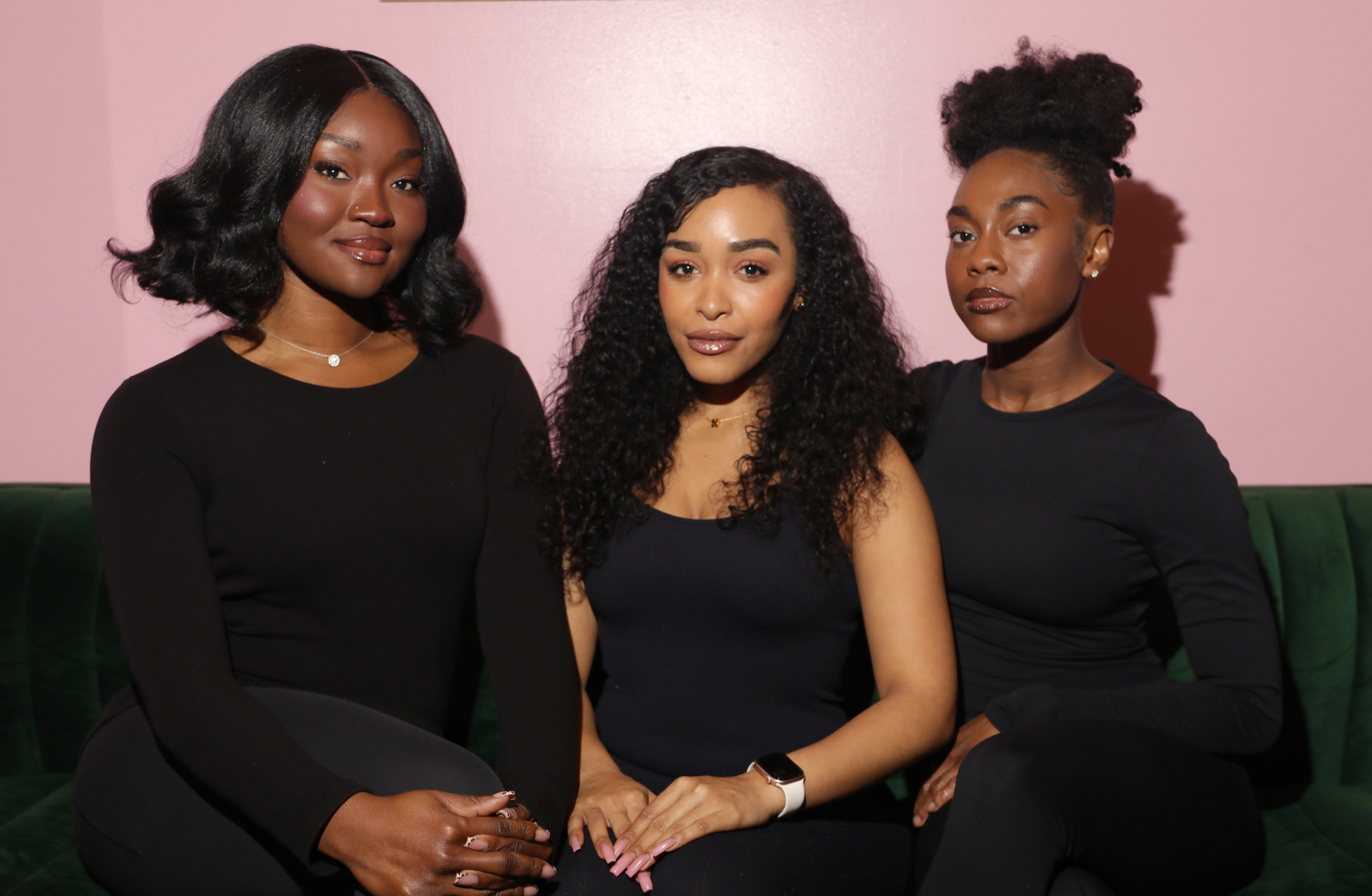 Three women sitting on a green couch against a pink wall, wearing black tops, posing for a photo.