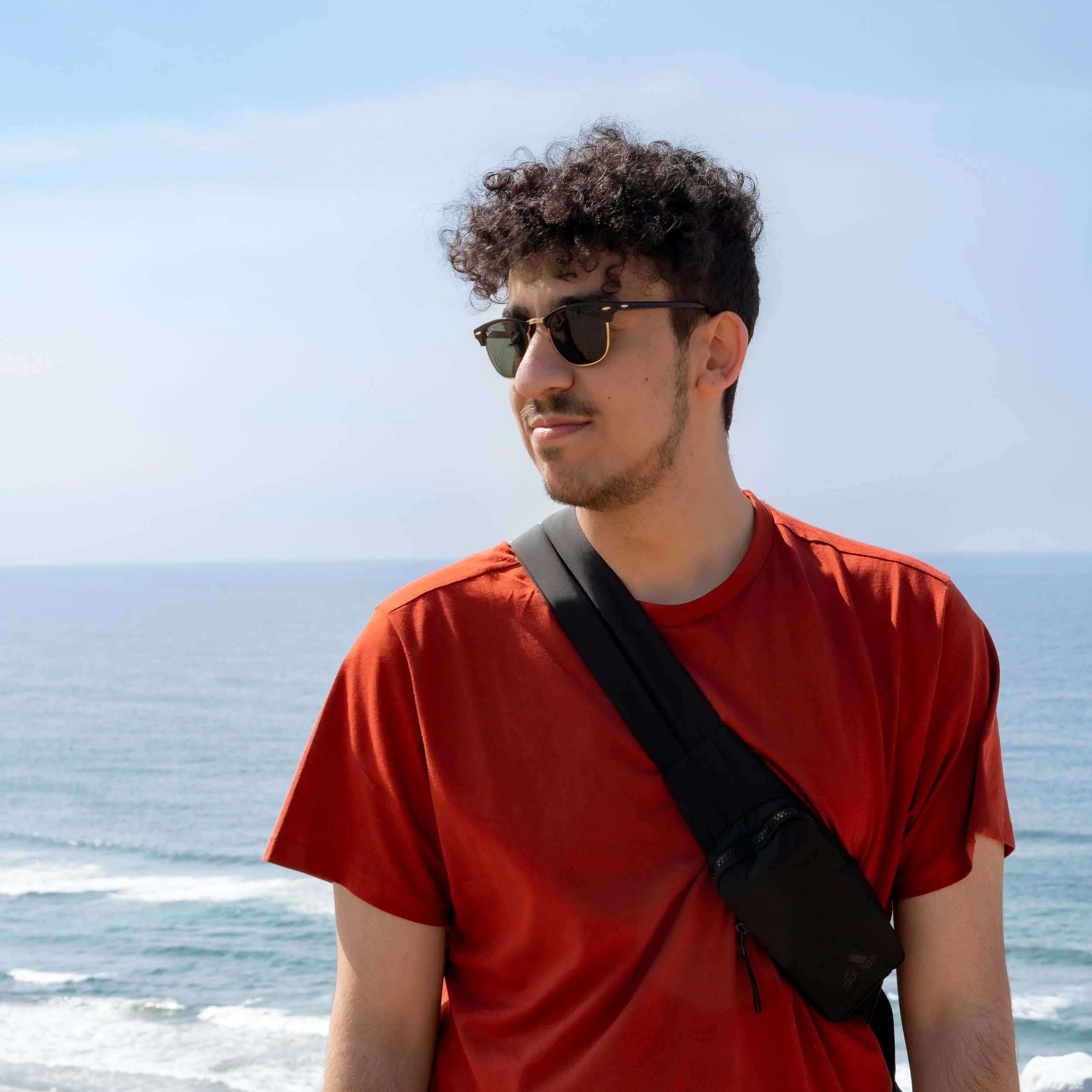 A young man with curly hair wearing sunglasses and a red t-shirt, standing by the ocean with waves in the background.