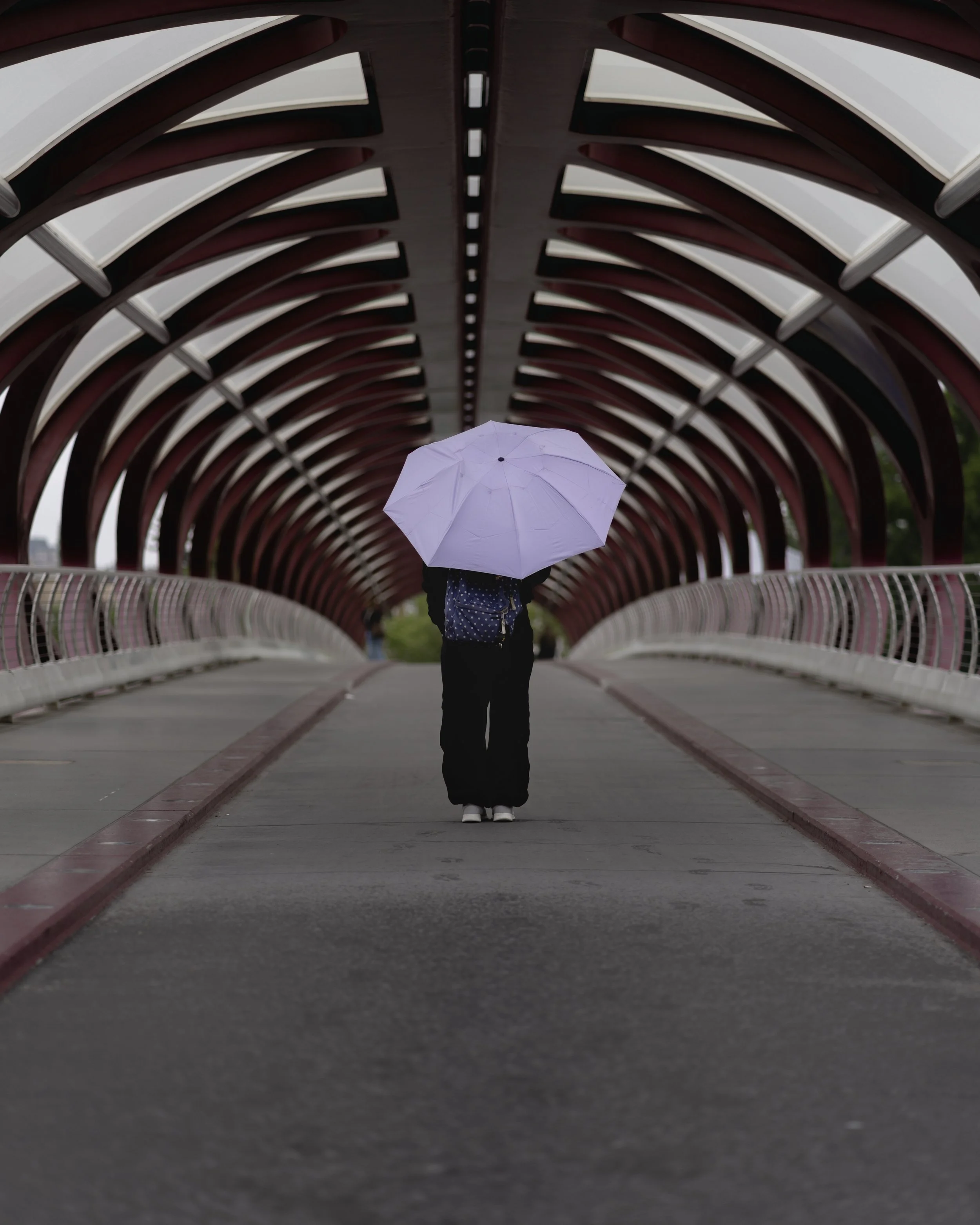 Person standing under a purple umbrella on a modern, arched pedestrian bridge