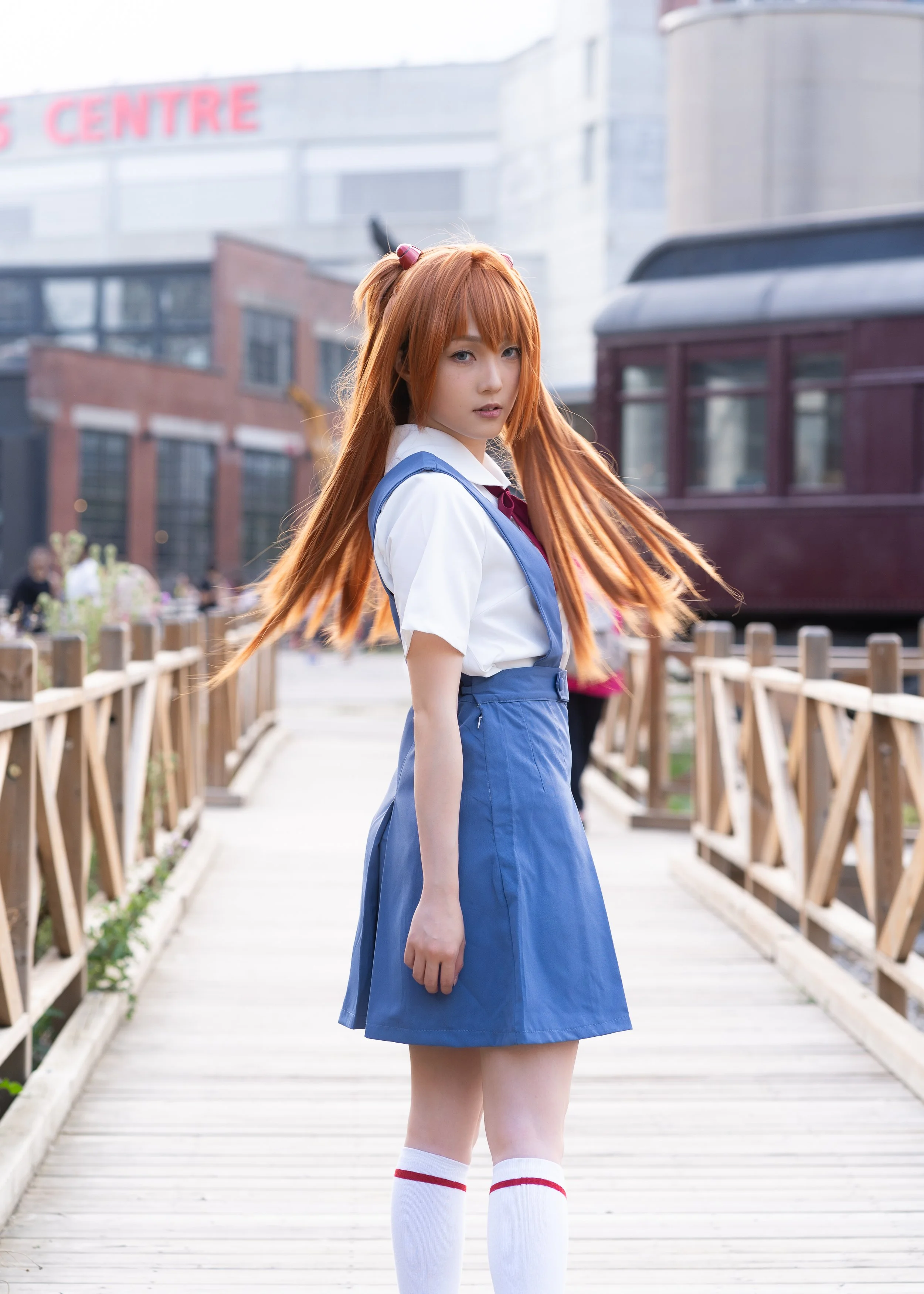 Young woman with long, orange hair wearing a Japanese school uniform standing on a wooden bridge outdoors with city buildings and a tram in the background.