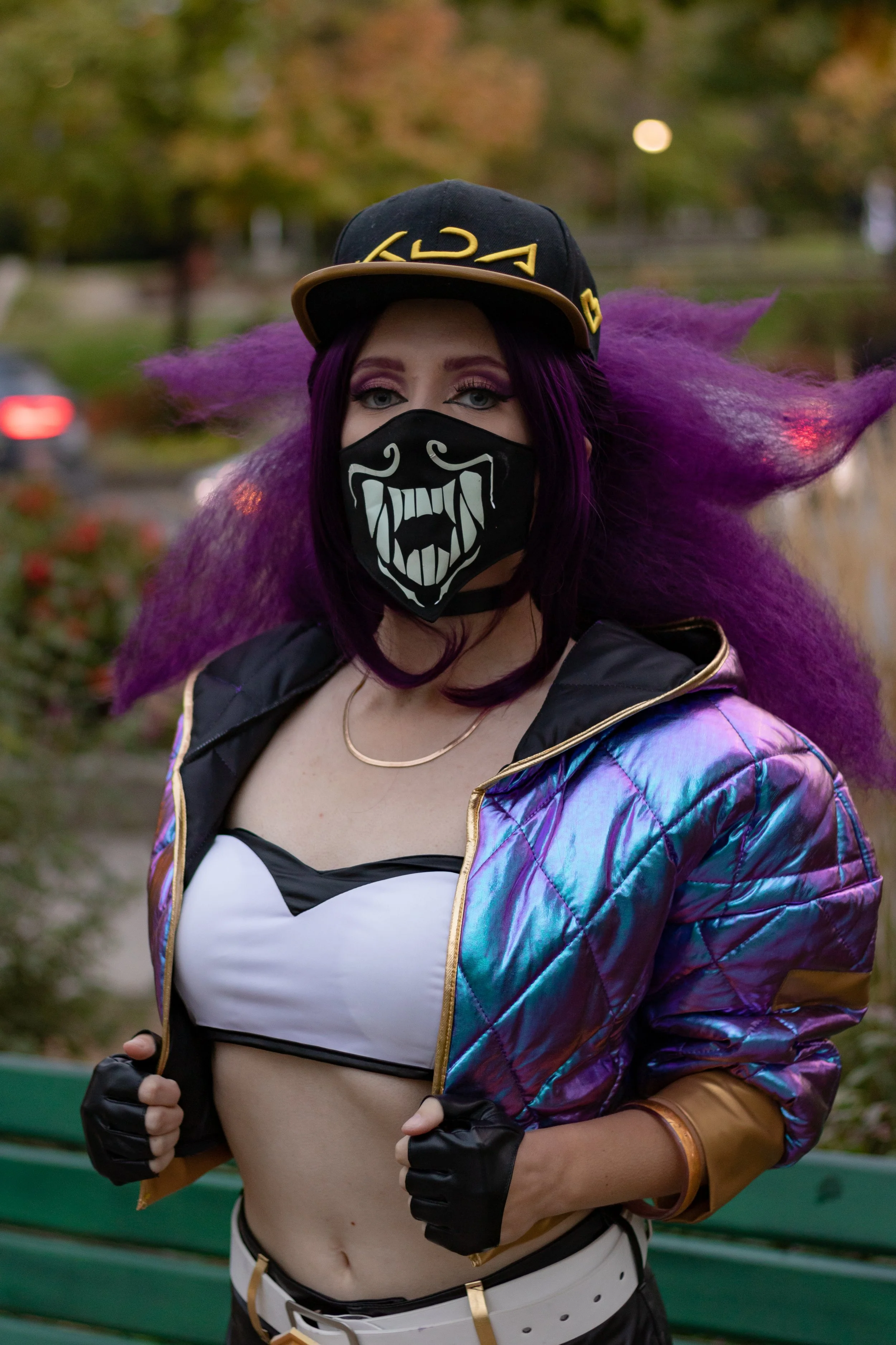 A woman with purple hair and makeup wearing a black cap, face mask with a white menacing face design, crop top, holographic jacket, and black gloves outdoors in a park.