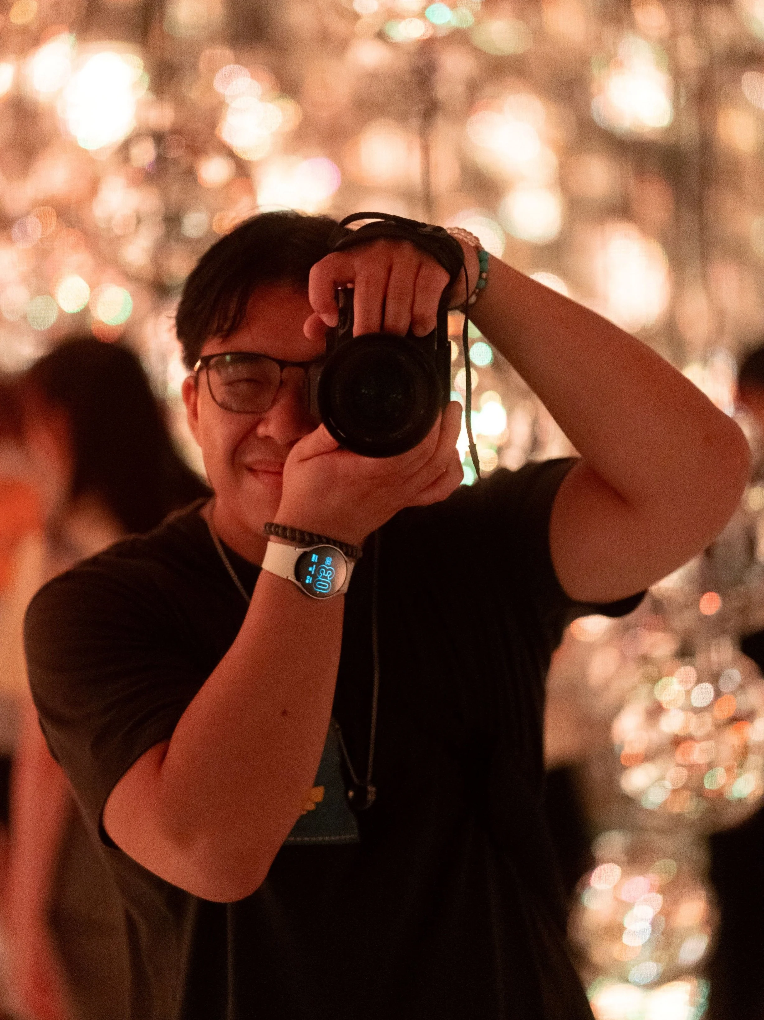 A person taking a selfie with a camera, smiling, in a festive setting with sparkling lights in the background.