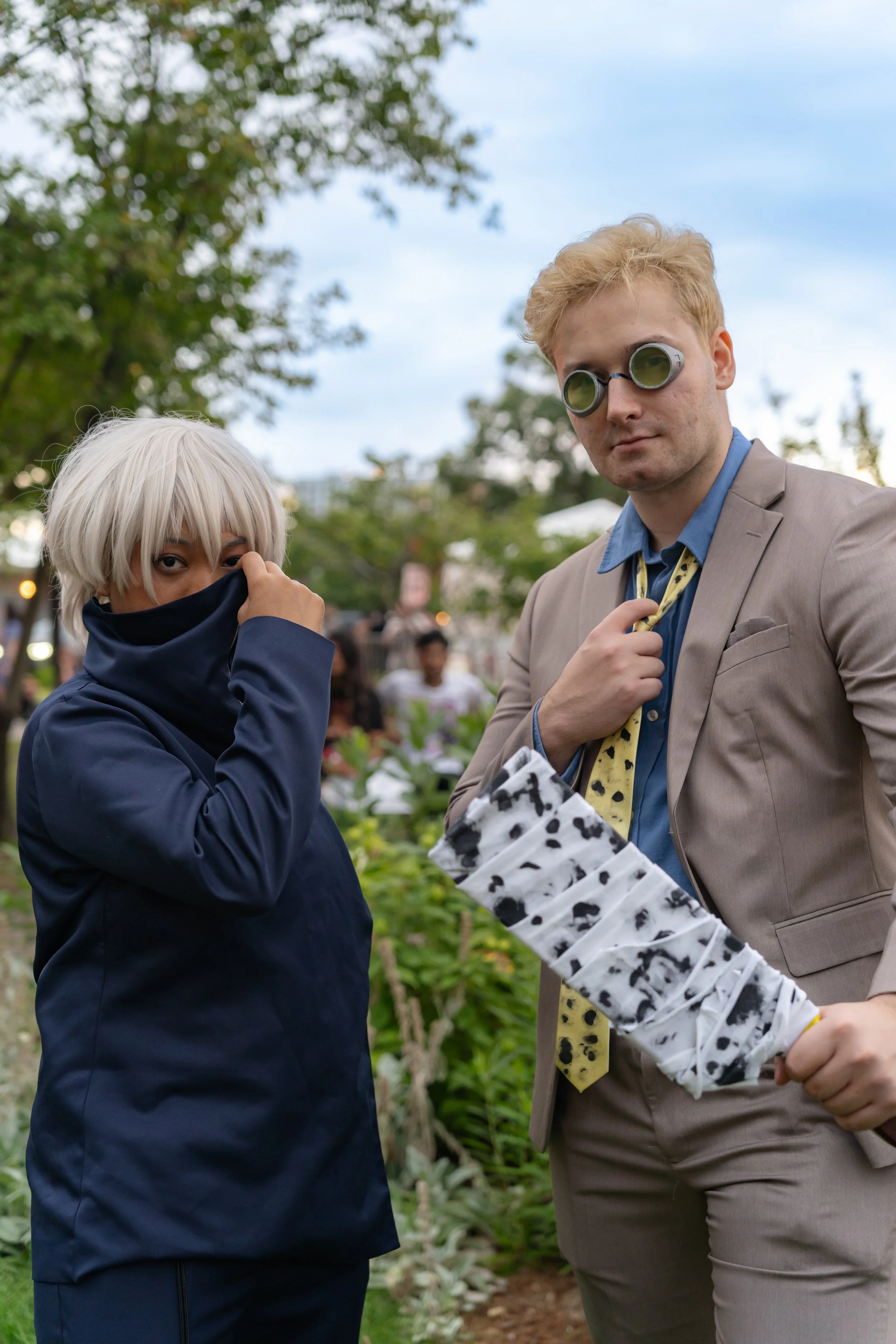 Two people in costumes at an outdoor event: one with silver hair covering their face with a dark blue jacket, and the other dressed as an eccentric businessman with sunglasses, a beige suit, and a yellow tie with black spots, holding a cow-print umbrella.