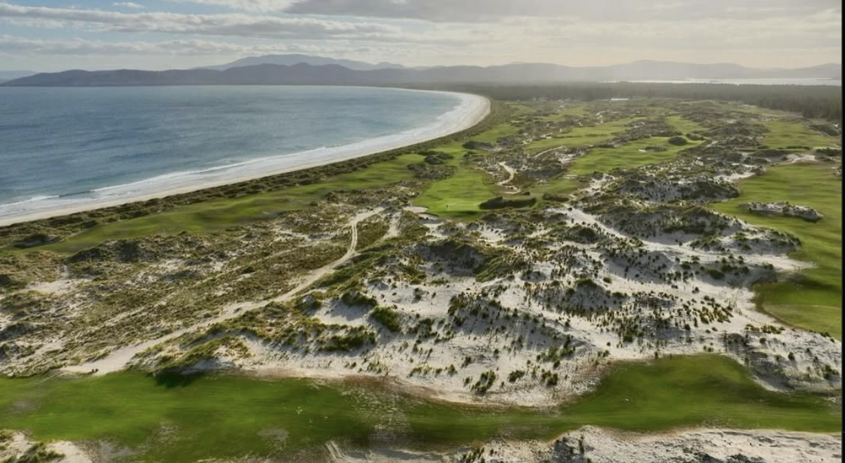 Aerial view of a coastal landscape featuring sandy dunes, green grass, and a beach, with ocean waves in the background and distant mountains under partly cloudy skies.