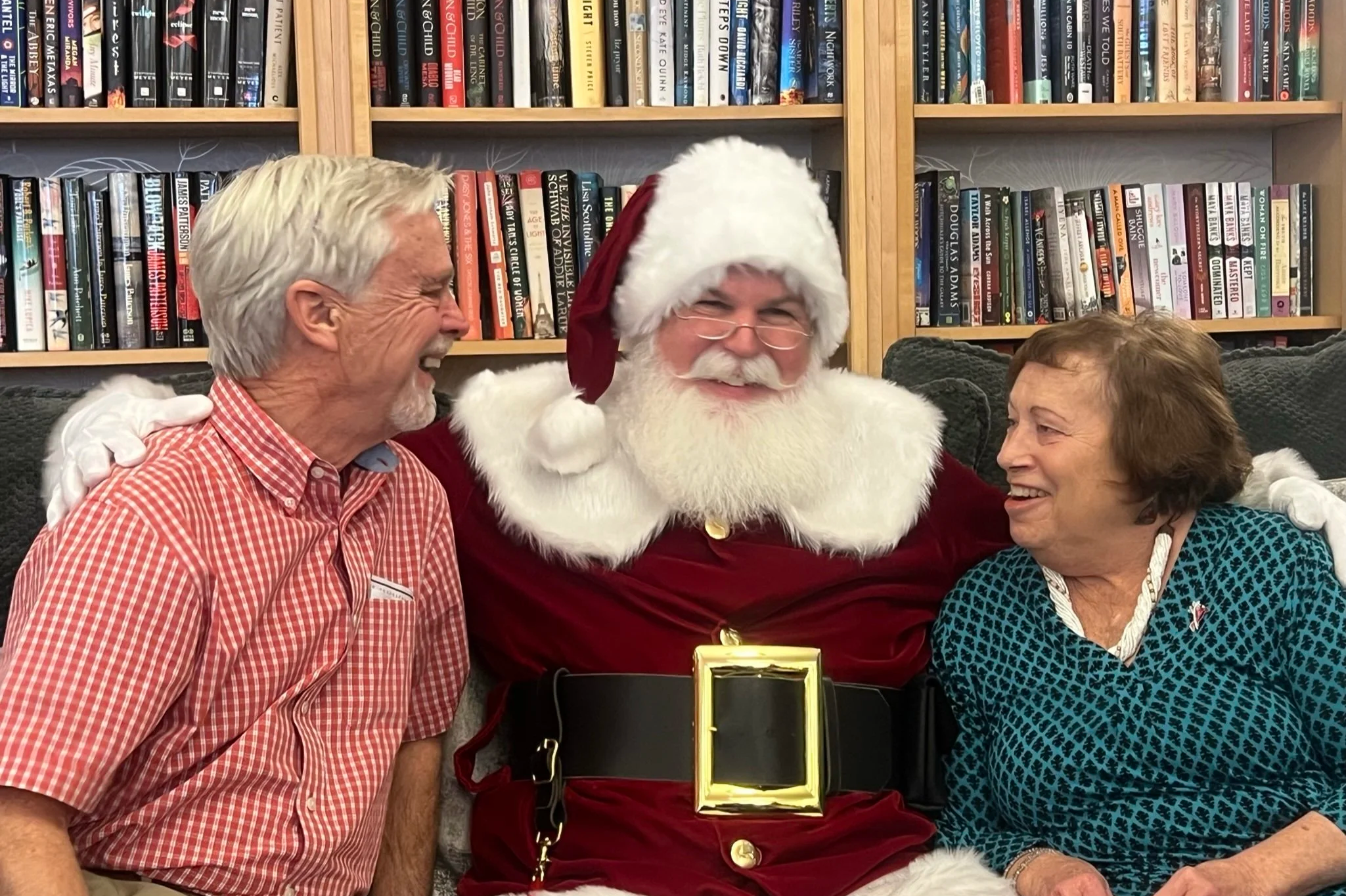People smiling with a person dressed as Santa Claus in a red suit, sitting in front of a bookshelf filled with books.