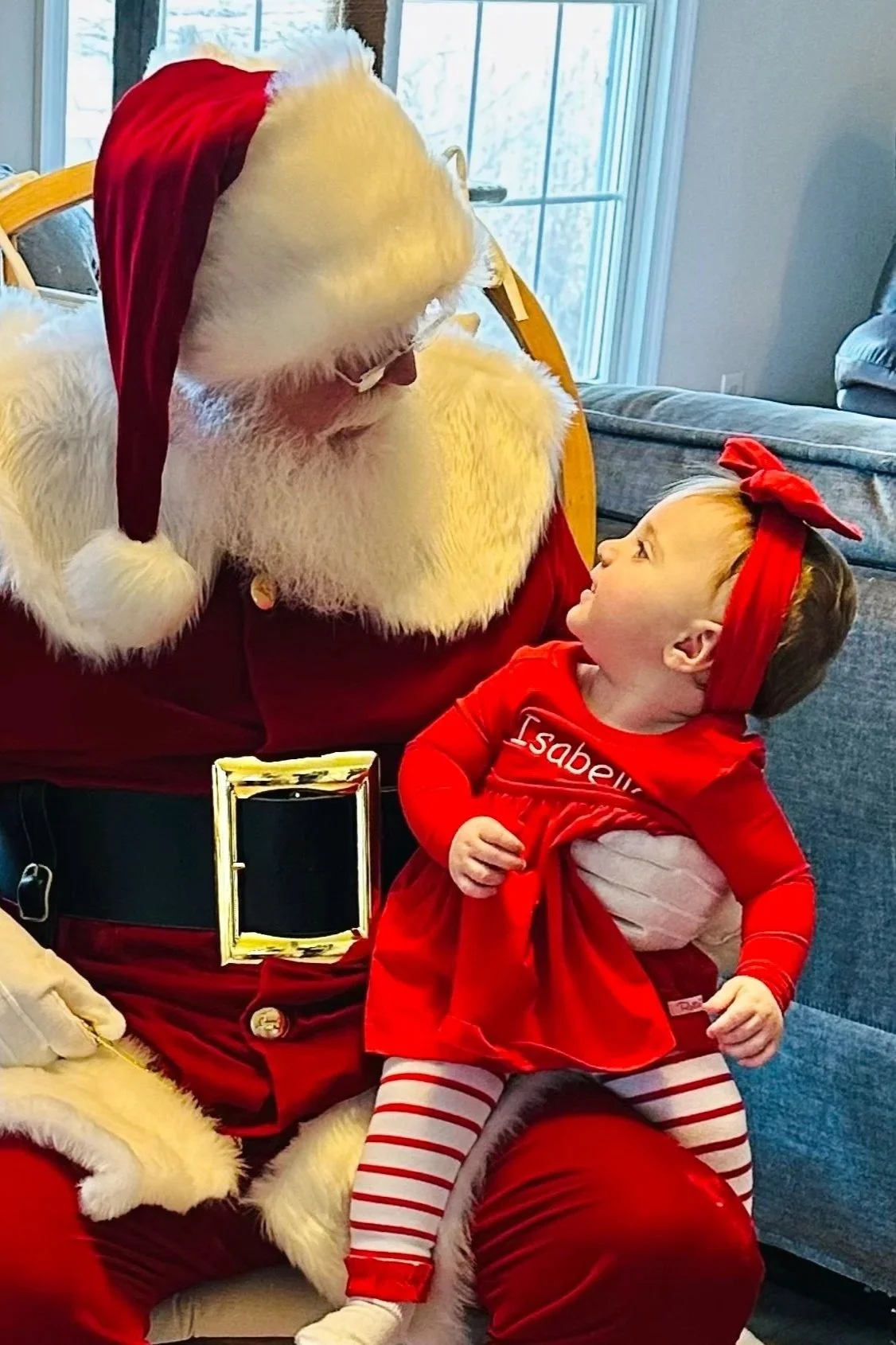 Santa Claus holding a baby in a red outfit indoors, next to a decorated railing and Christmas tree.