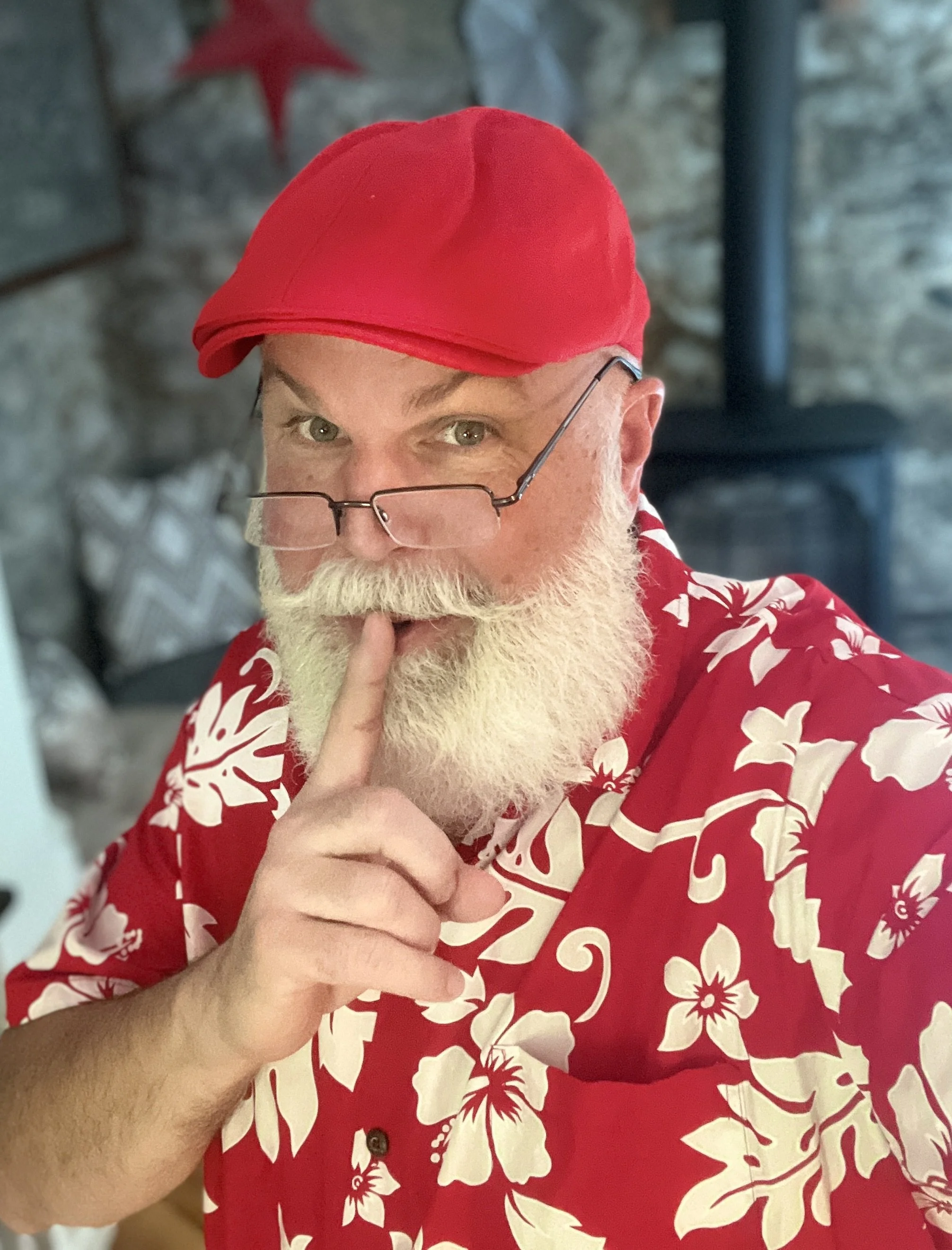 Man with a white beard wearing a red cap and Hawaiian shirt, holding a finger to his lips in a shushing gesture.