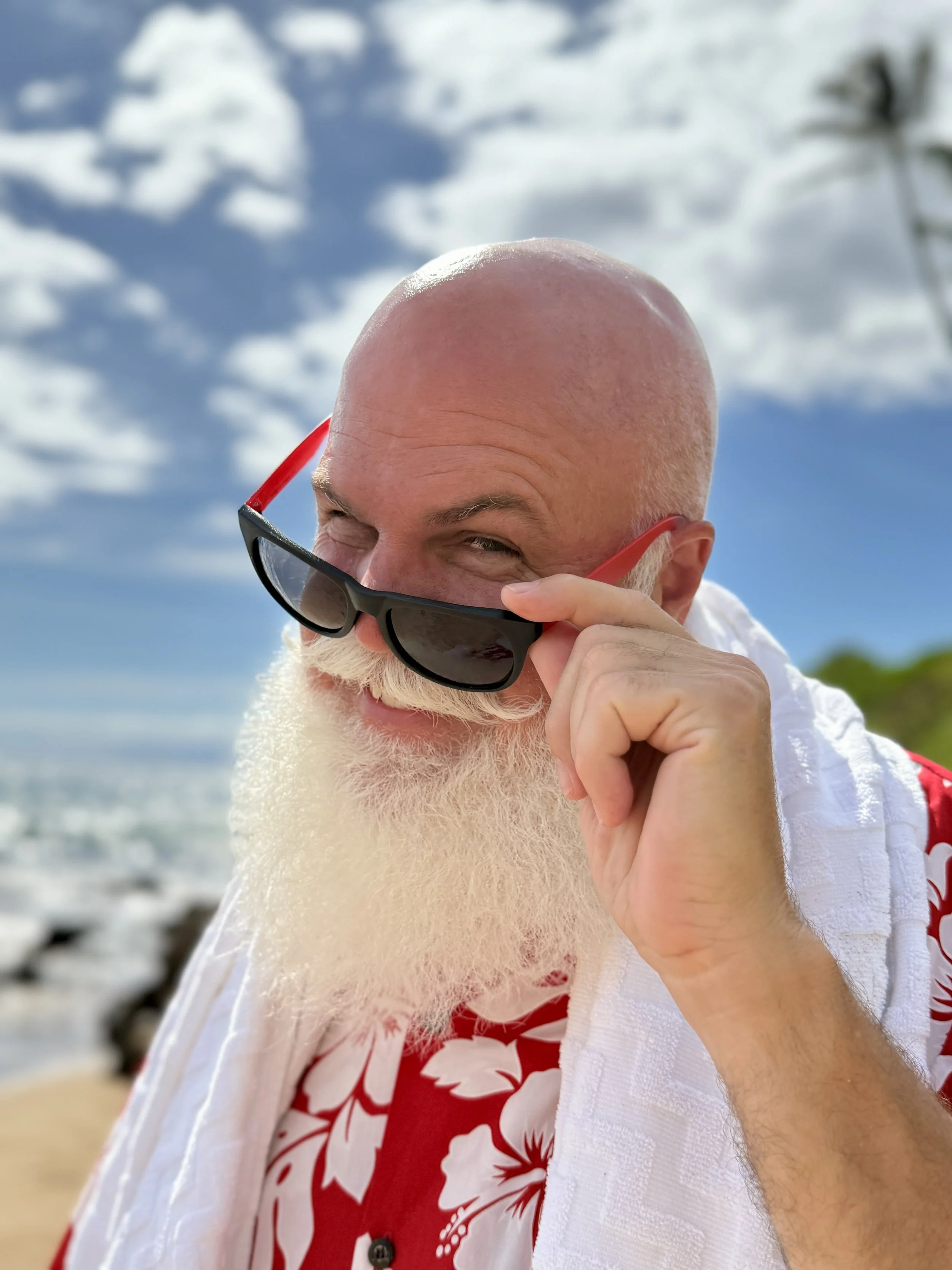 A man with a large white beard and a bald head, wearing a red and white floral shirt and holding sunglasses, smiling at the camera on a beach during daytime.