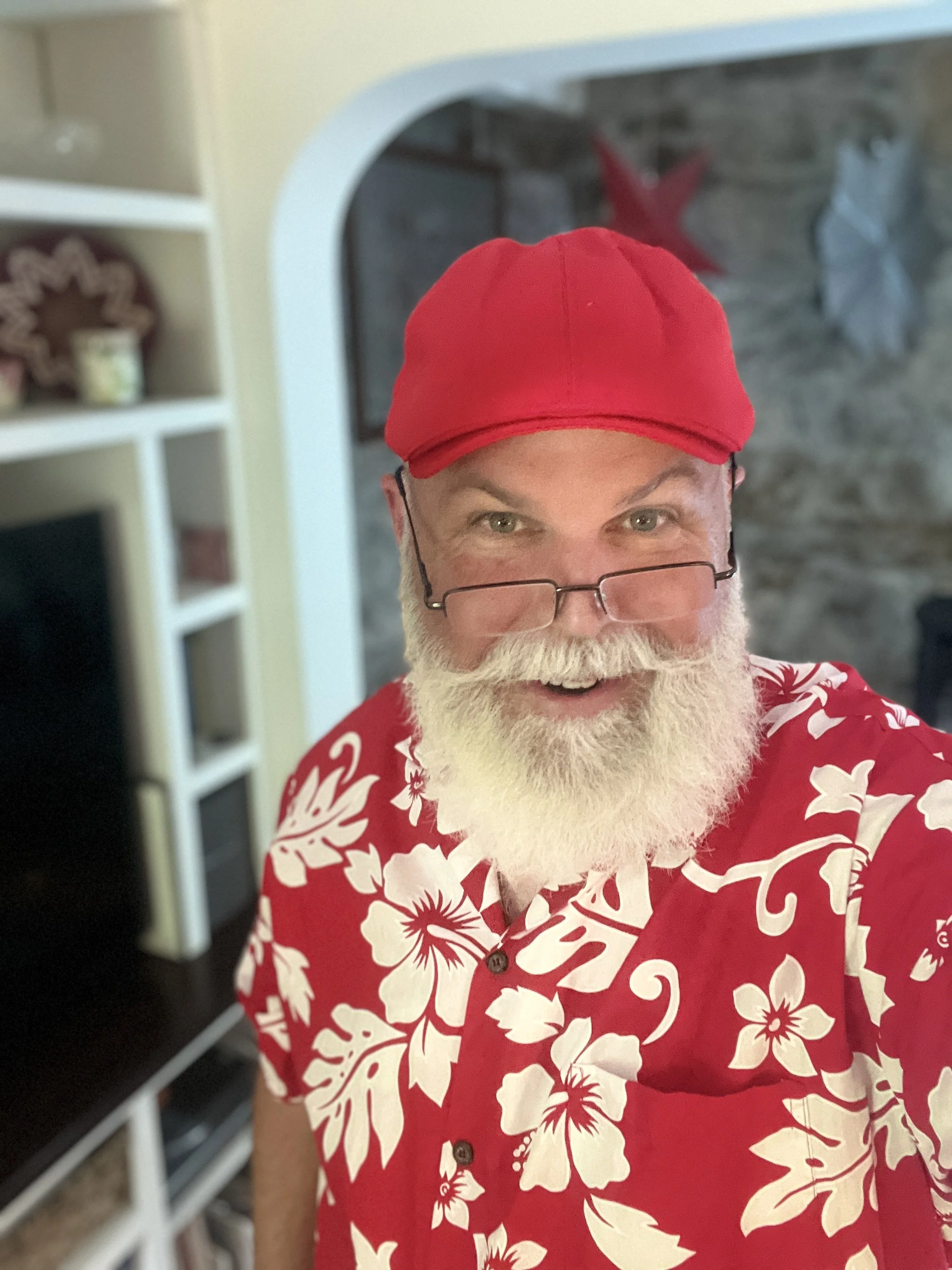 Man with white beard, wearing red floral shirt and red cap, standing indoors.