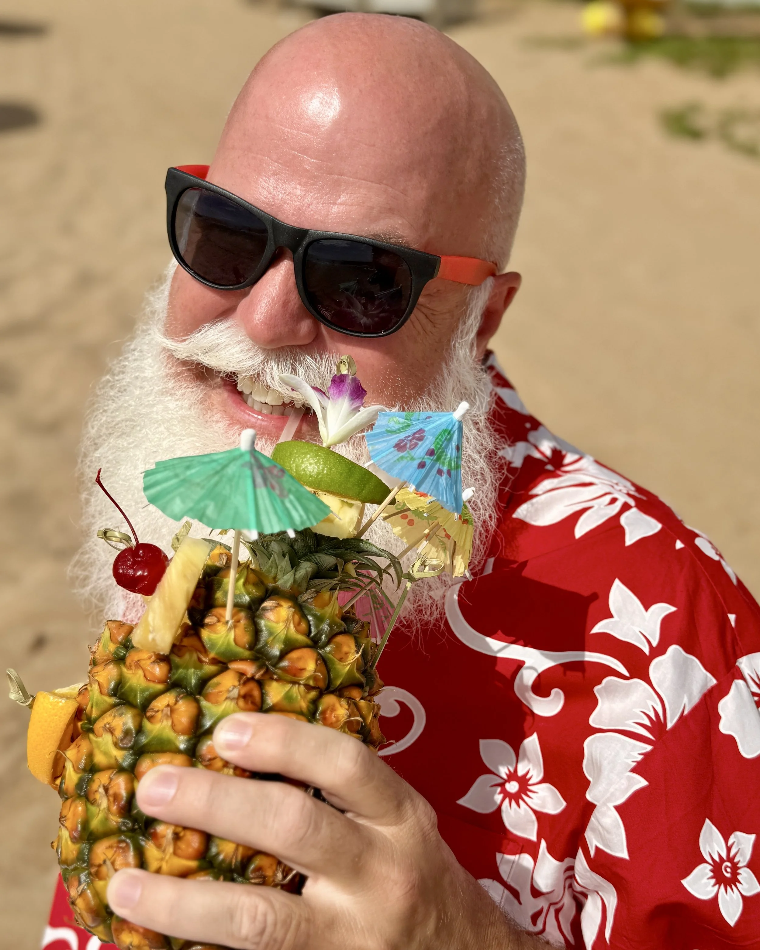 A man with a bald head and white beard wearing sunglasses and a red Hawaiian shirt is smiling while holding a pineapple decorated with colorful paper umbrellas, flowers, and fruit slices on a sandy beach.