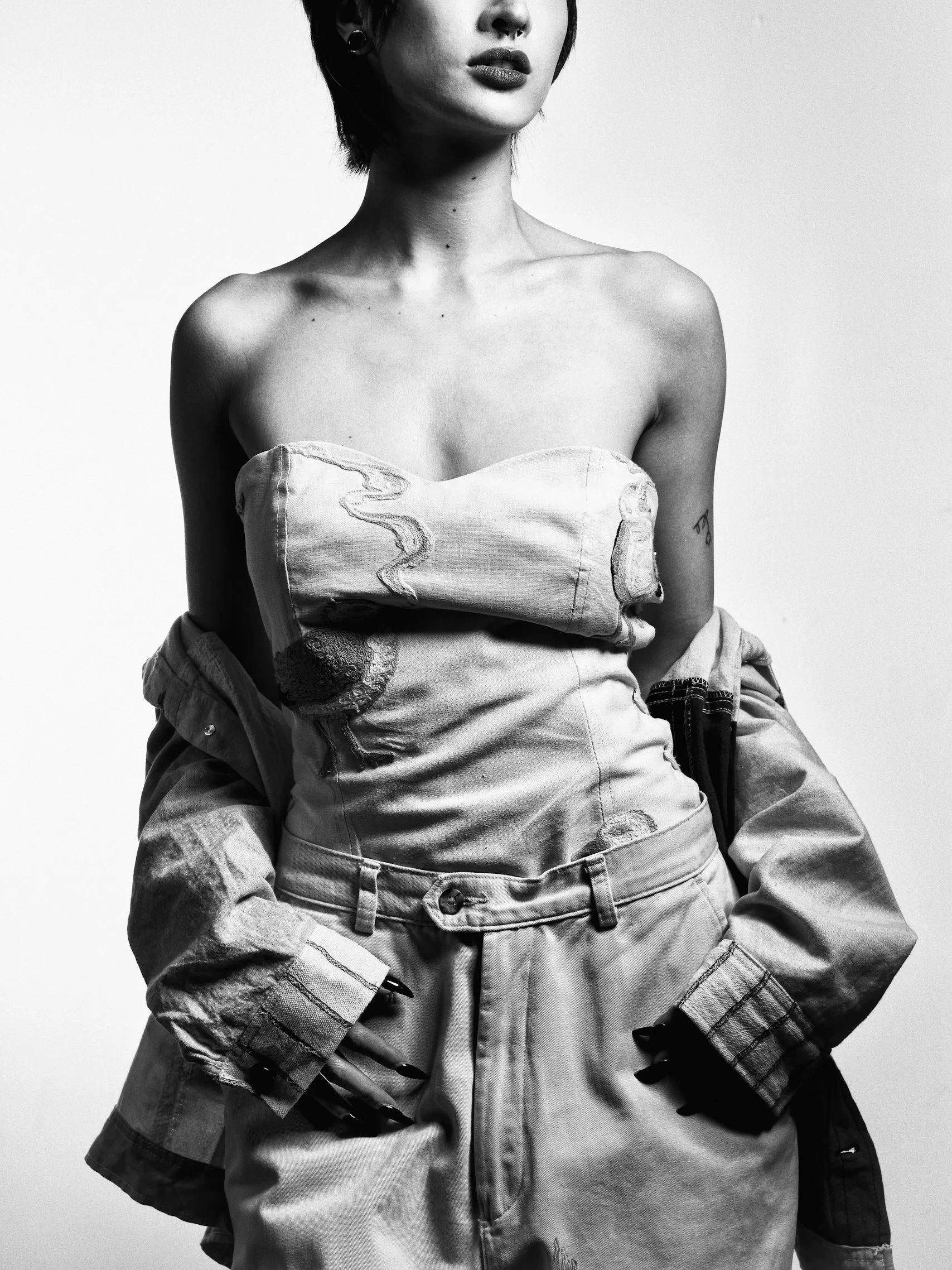 Black and white photo of a woman with short hair, wearing a strapless top, an oversized jacket, and baggy jeans. Her hands are in her pockets and she has dark nail polish.